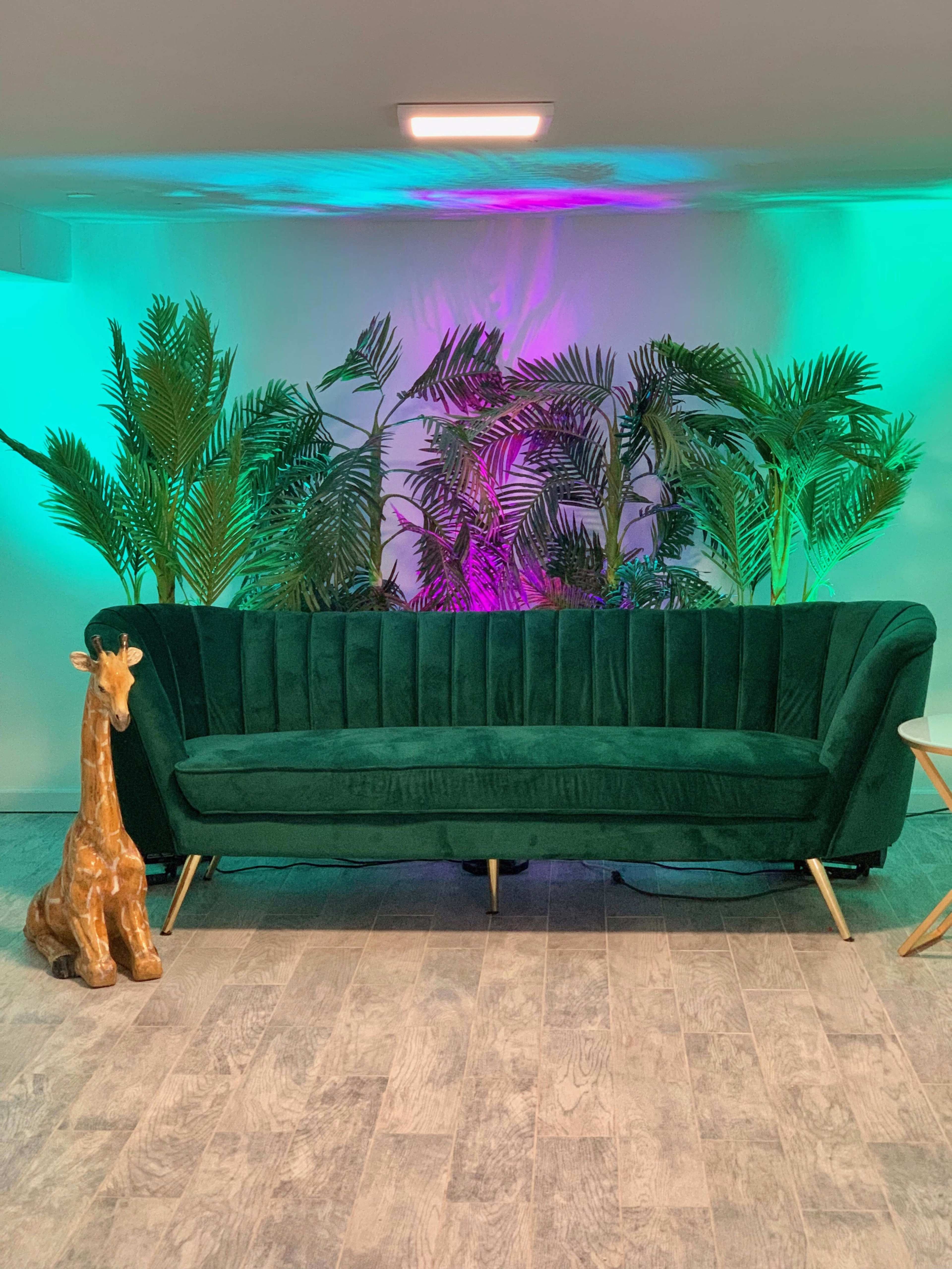 A green velvet sofa sits in front of a colorful backdrop of palm plants under a warm light.