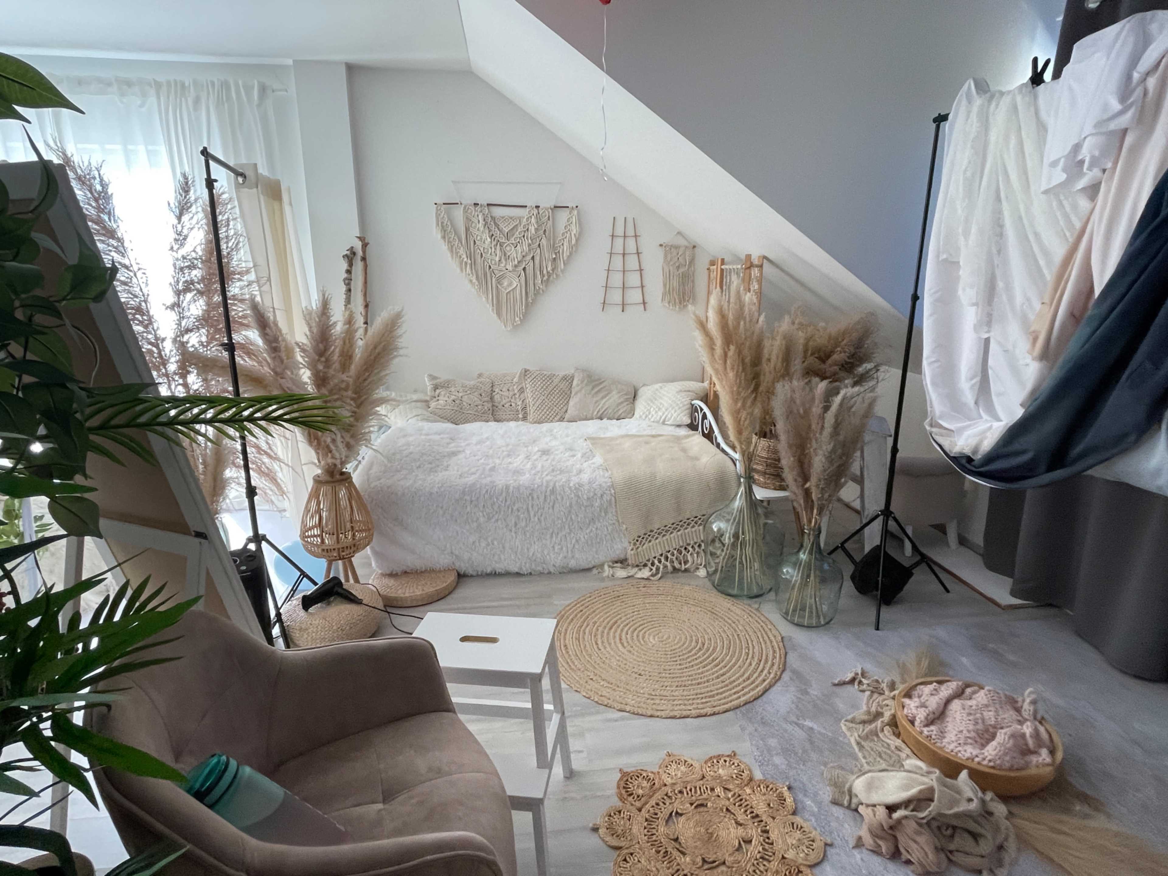 The image shows a cozy bedroom with a large bed adorned with light-colored blankets, surrounded by decorative plants and textiles.