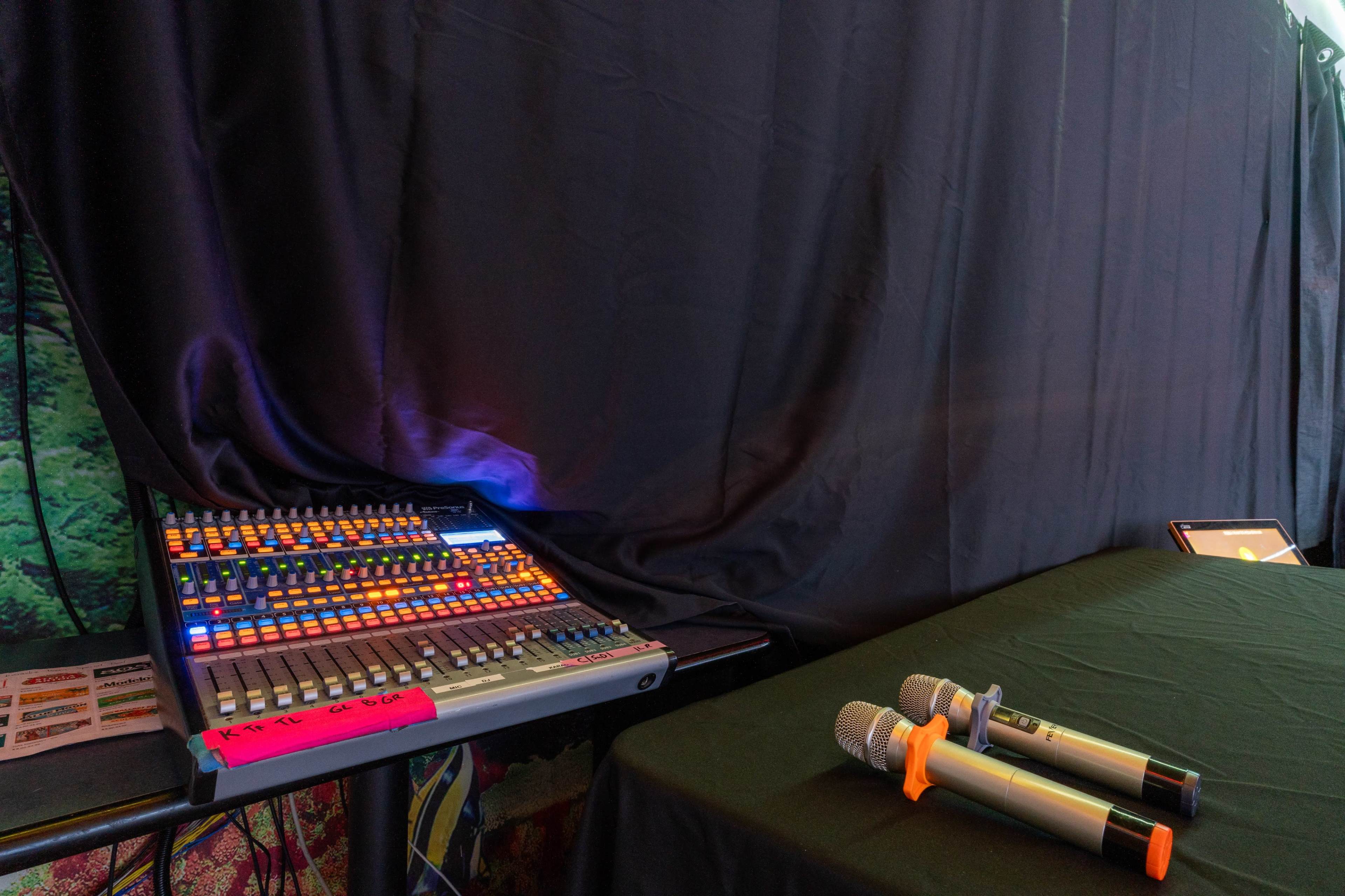 A sound mixing console with colorful controls sits on a table beside two microphones, against a backdrop of a black curtain.