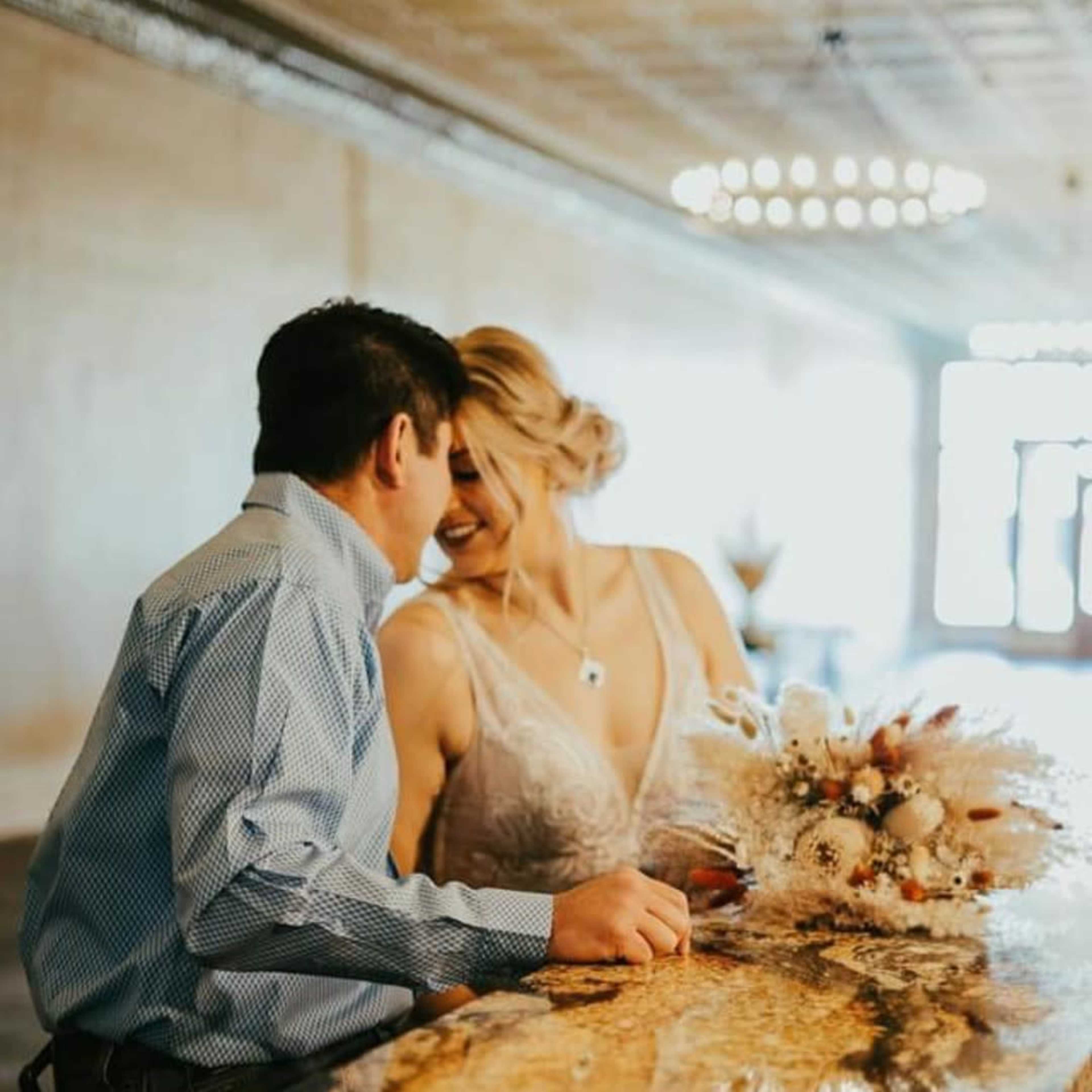 A couple leans toward each other at a bar counter, with the woman holding a bouquet and wearing an elegant dress.