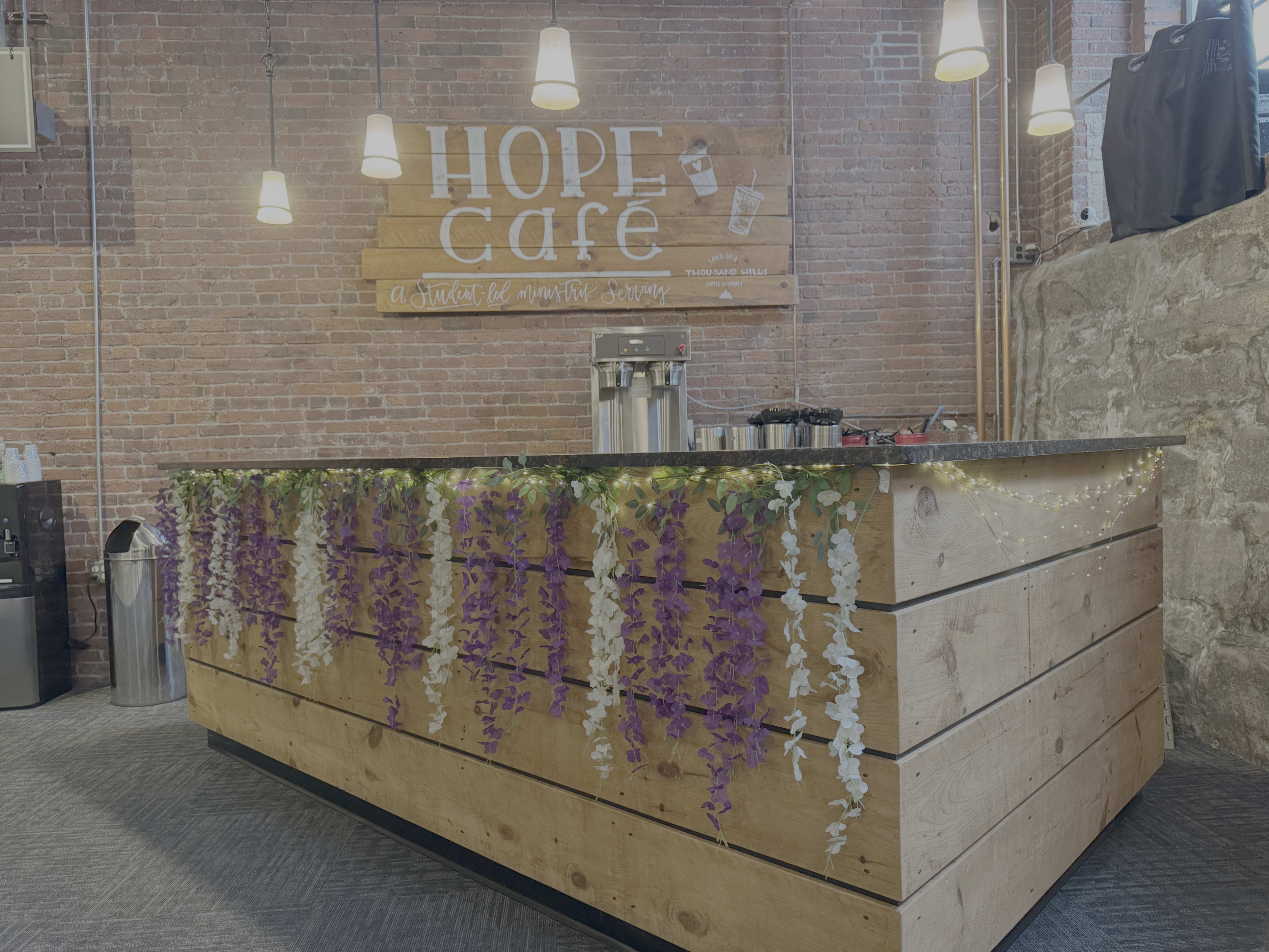 The image shows a wooden bar counter decorated with purple and white flowers, under hanging lights, against a backdrop of a brick wall with a "Hope Café" sign.
