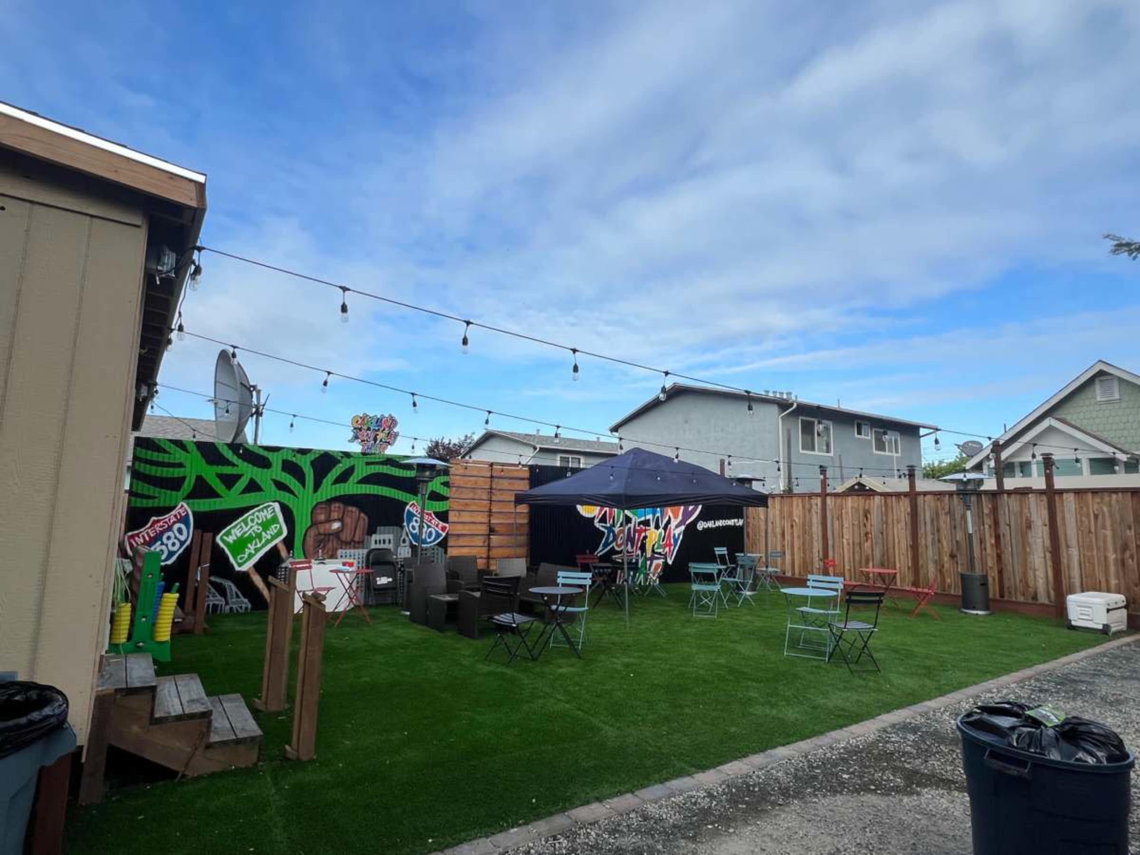 The image shows a fenced outdoor area with artificial grass, featuring several tables and chairs, a tent, and vibrant murals on a wall.