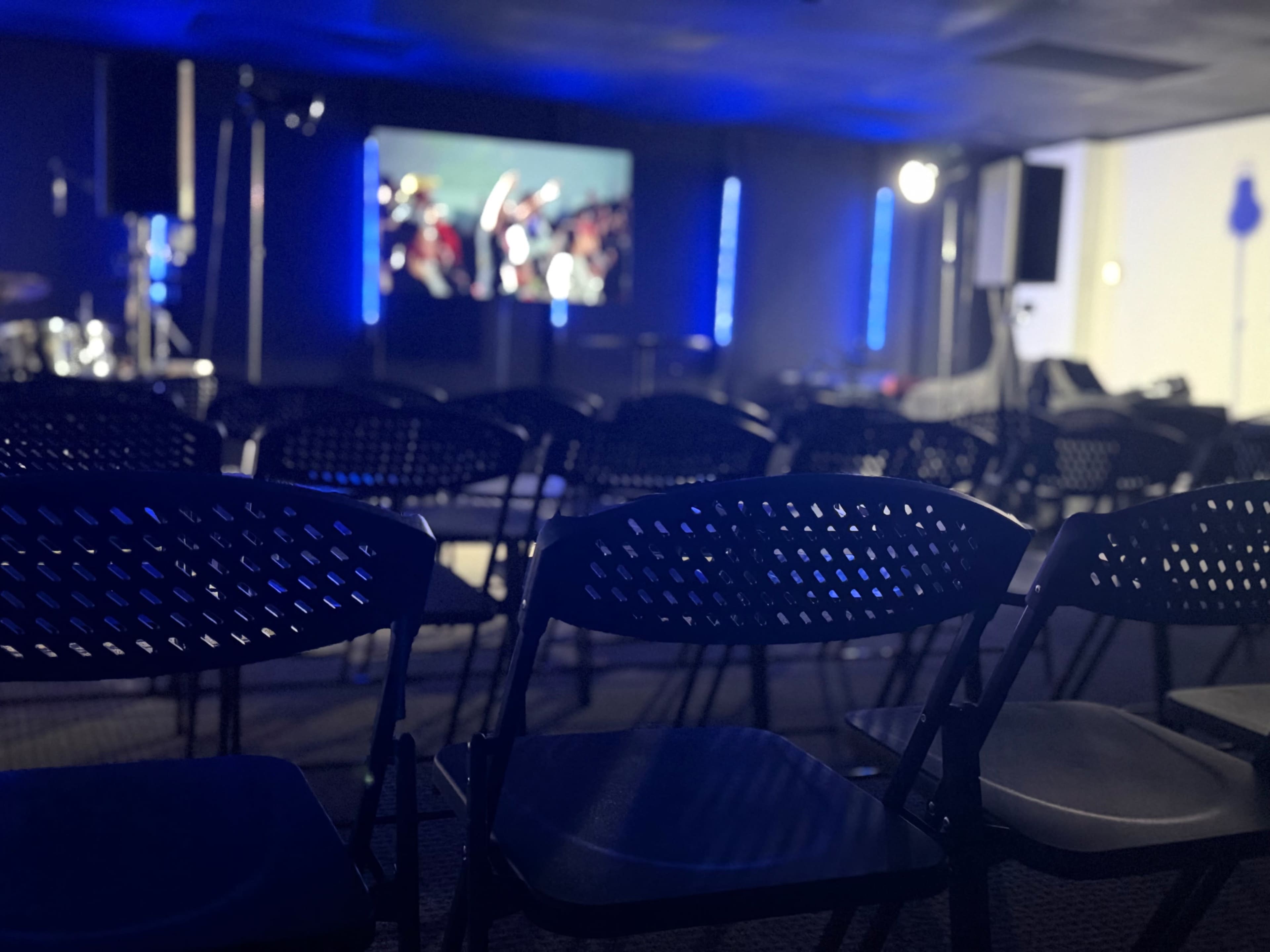 A conference room is set up with rows of black chairs facing a screen that displays a crowd, illuminated by blue lights.