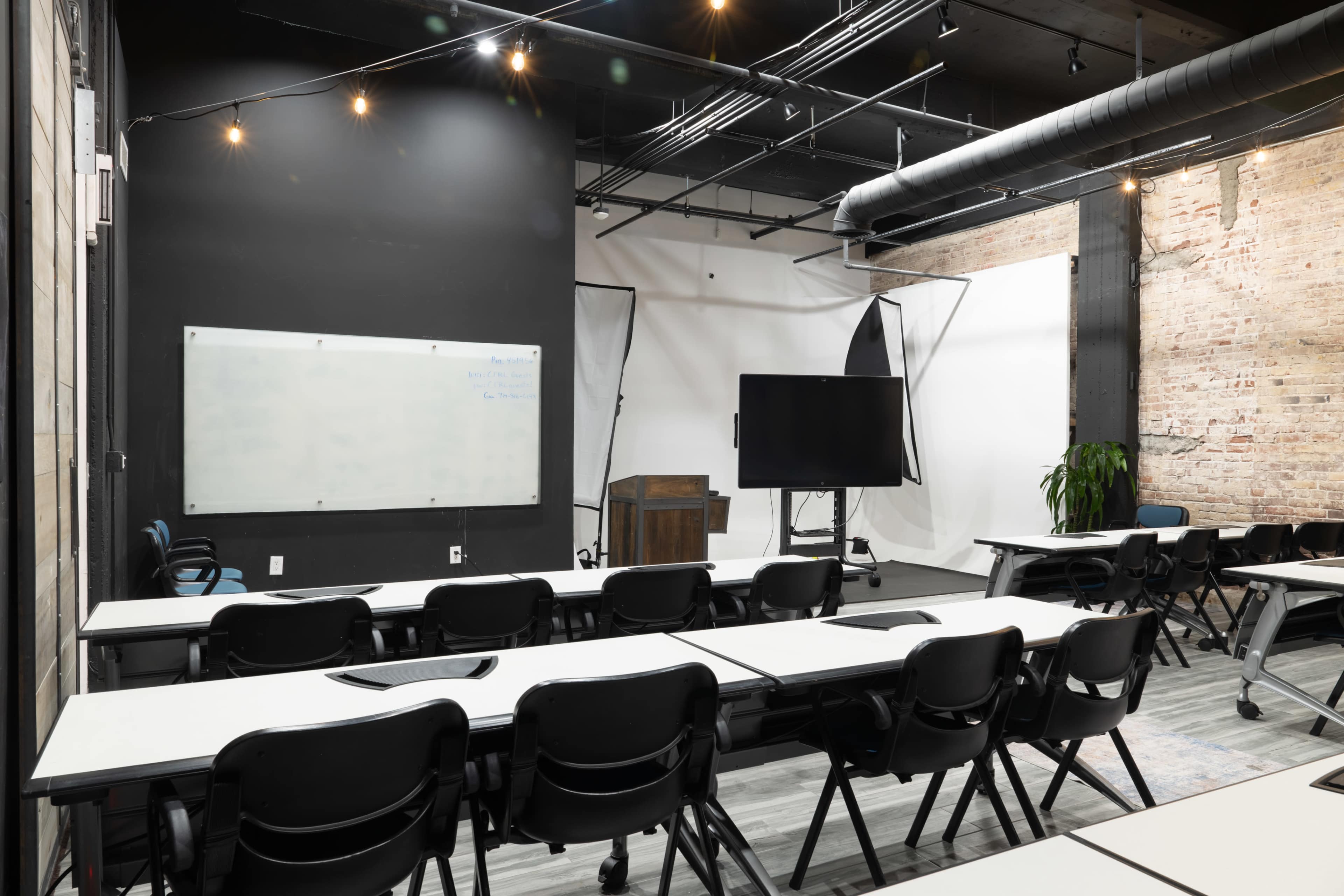 The image shows a modern conference room with several rows of tables and chairs, a whiteboard, and a large screen against a textured wall.