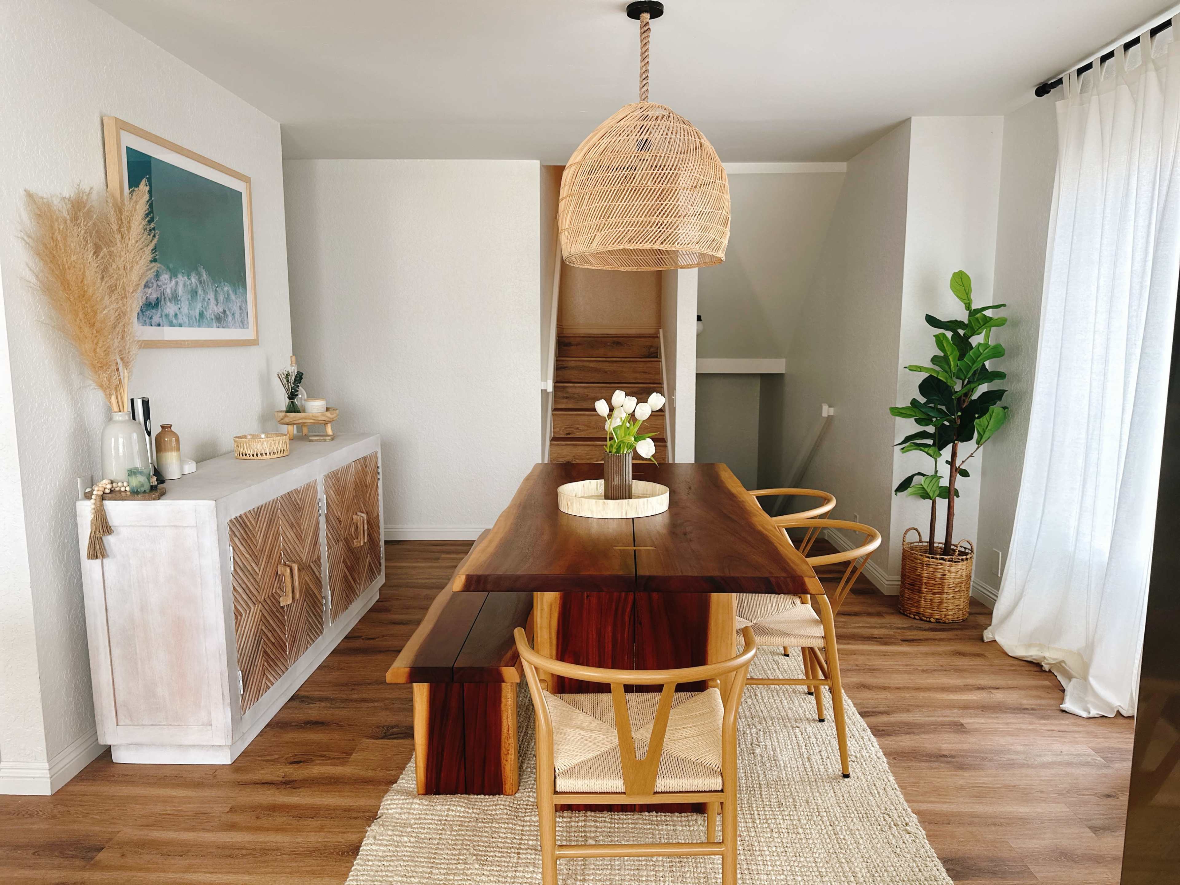 A modern dining area features a wooden table surrounded by chairs, a textured rug, a sideboard, and decorative plants.