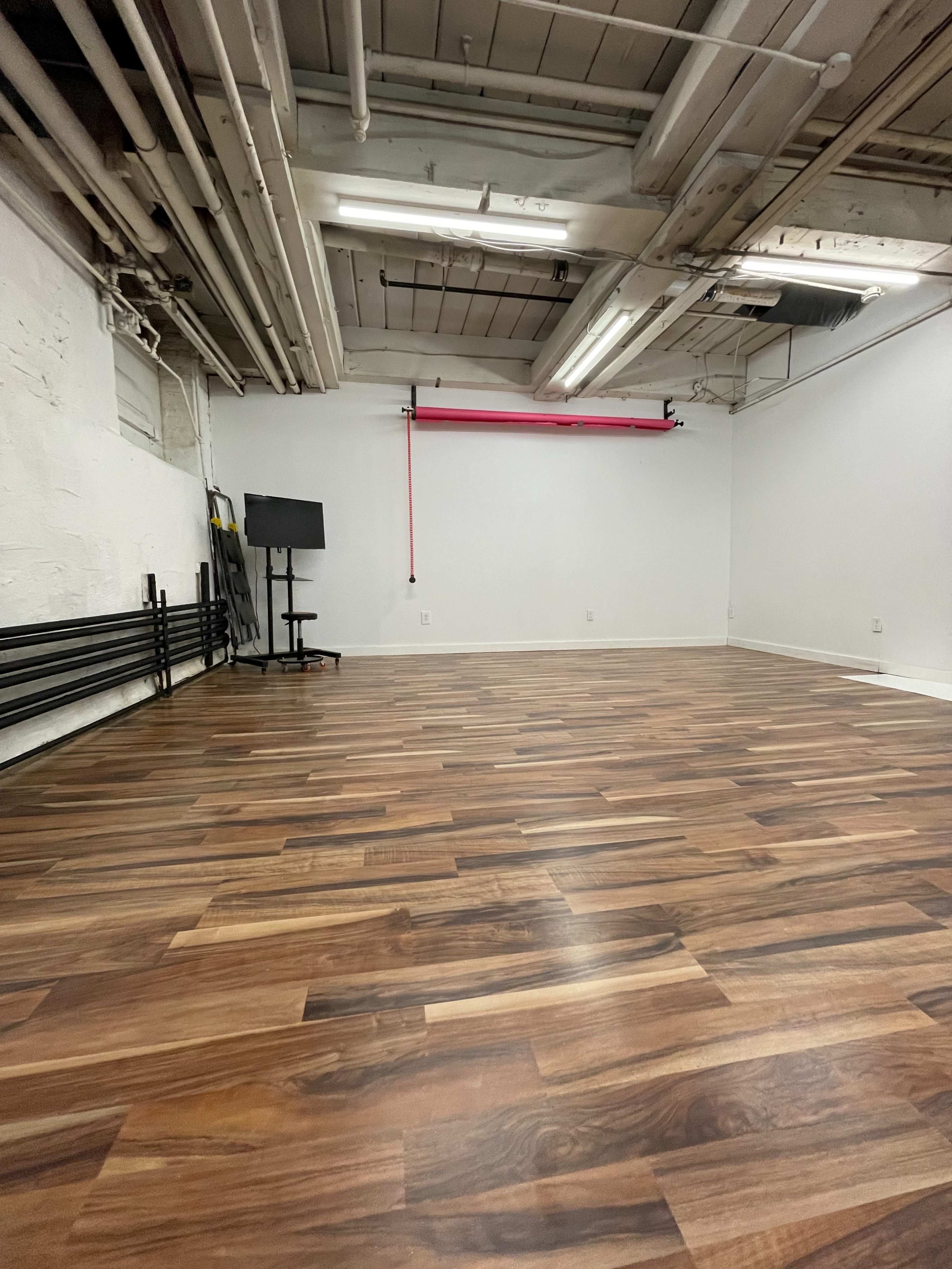An empty room with wooden flooring, exposed ceiling beams, and a wall-mounted red pipe, along with a black monitor stand in one corner.