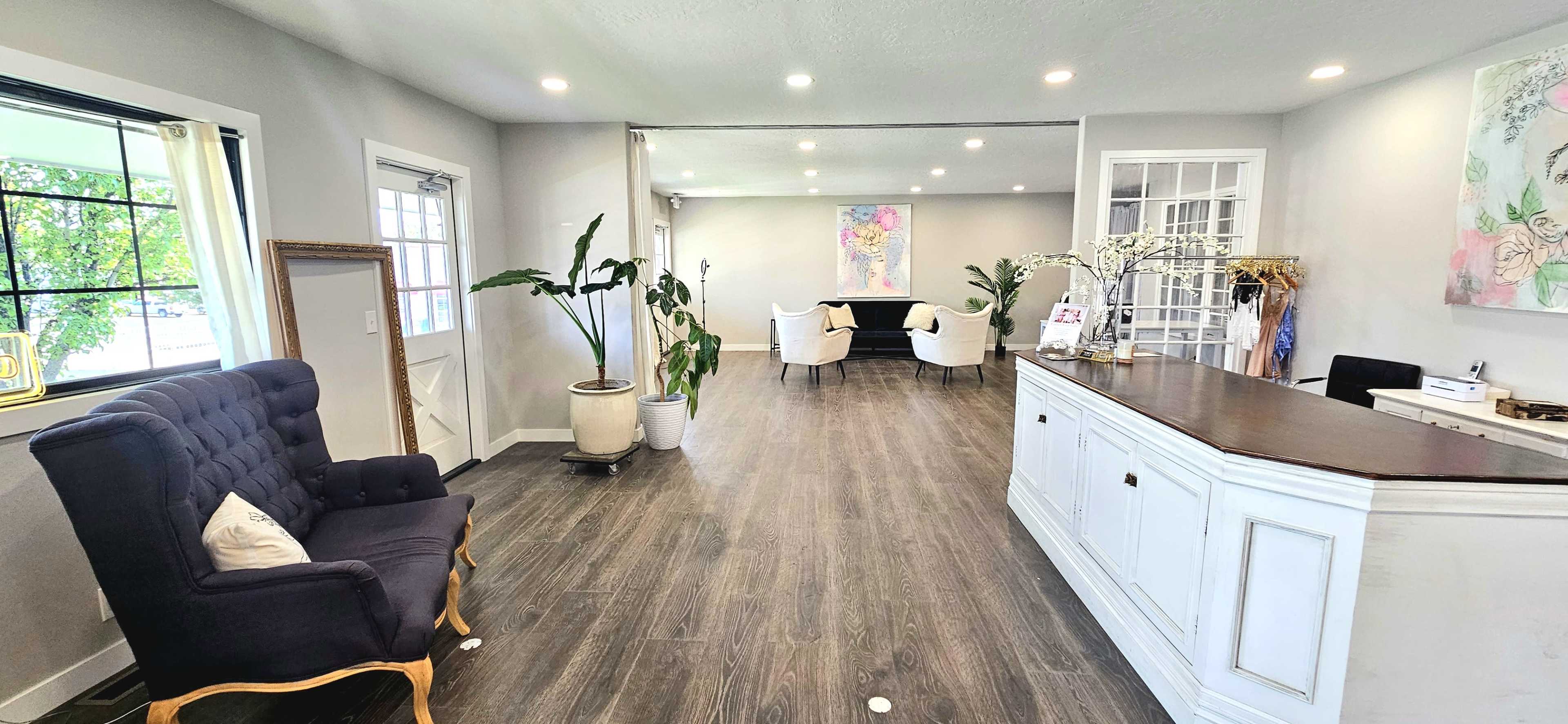 A spacious, well-lit reception area features a dark blue armchair, a white reception desk, and modern decor with plants and artwork against light gray walls.