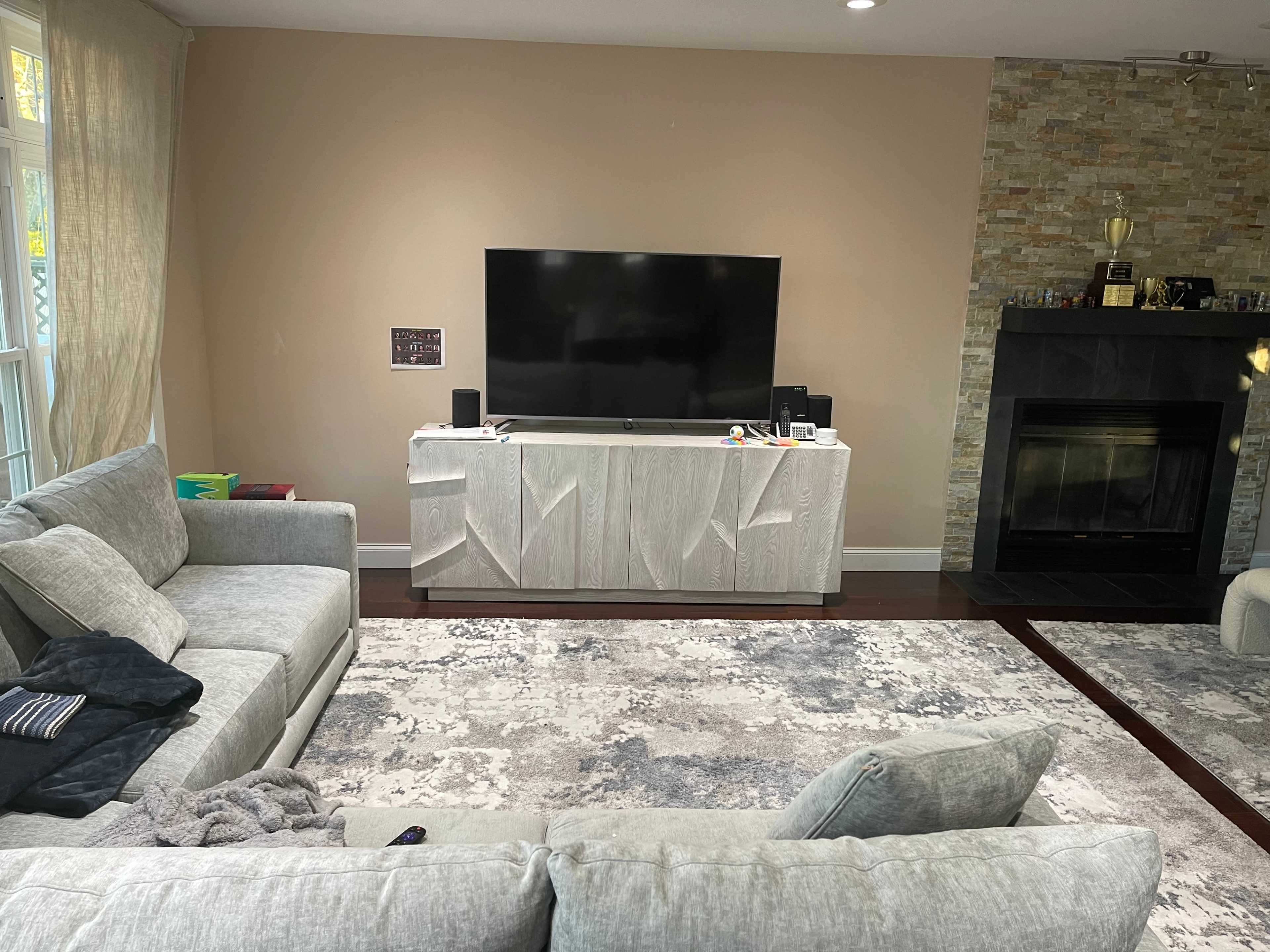 The image shows a living room with a television mounted above a light-colored cabinet, flanked by a stone fireplace and two grey sofas positioned around a patterned rug.