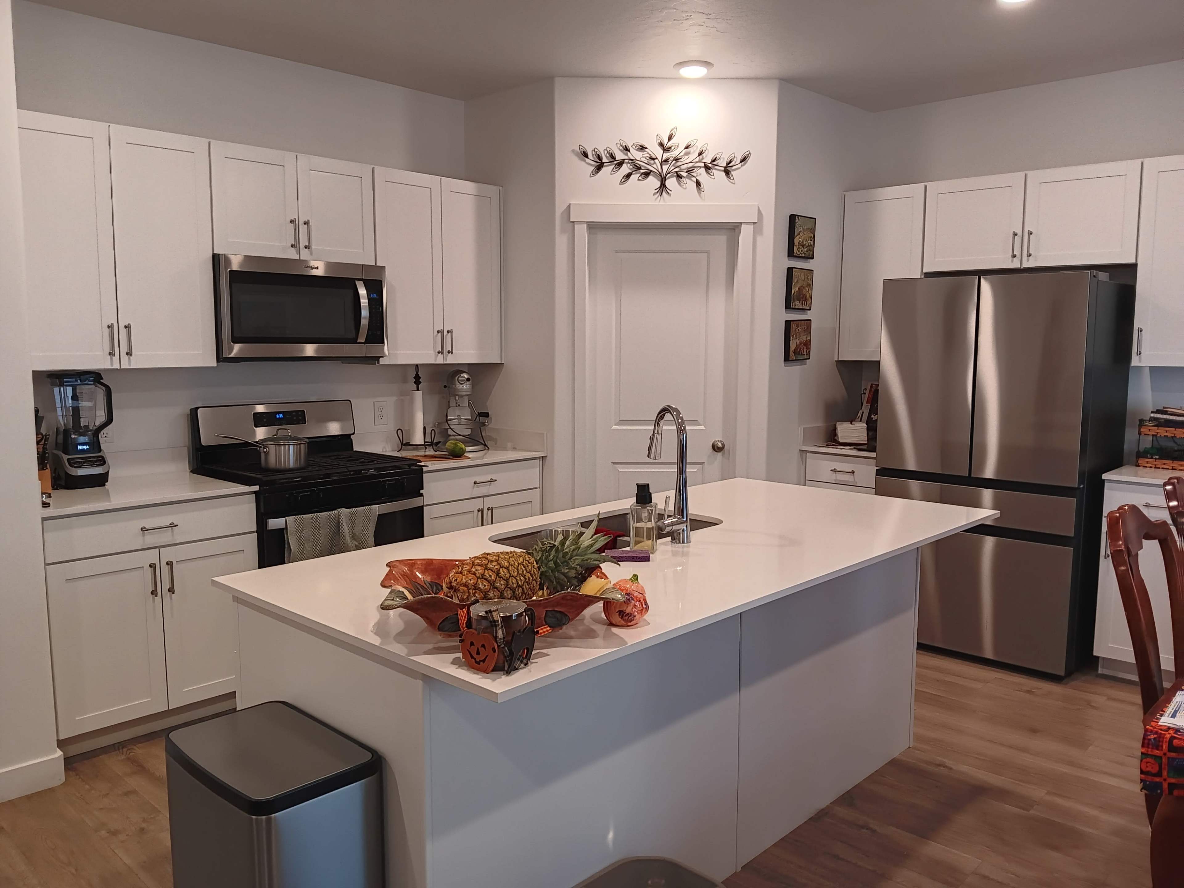 A modern kitchen features white cabinetry, stainless steel appliances, an island with a fruit display, and light-colored wooden flooring.