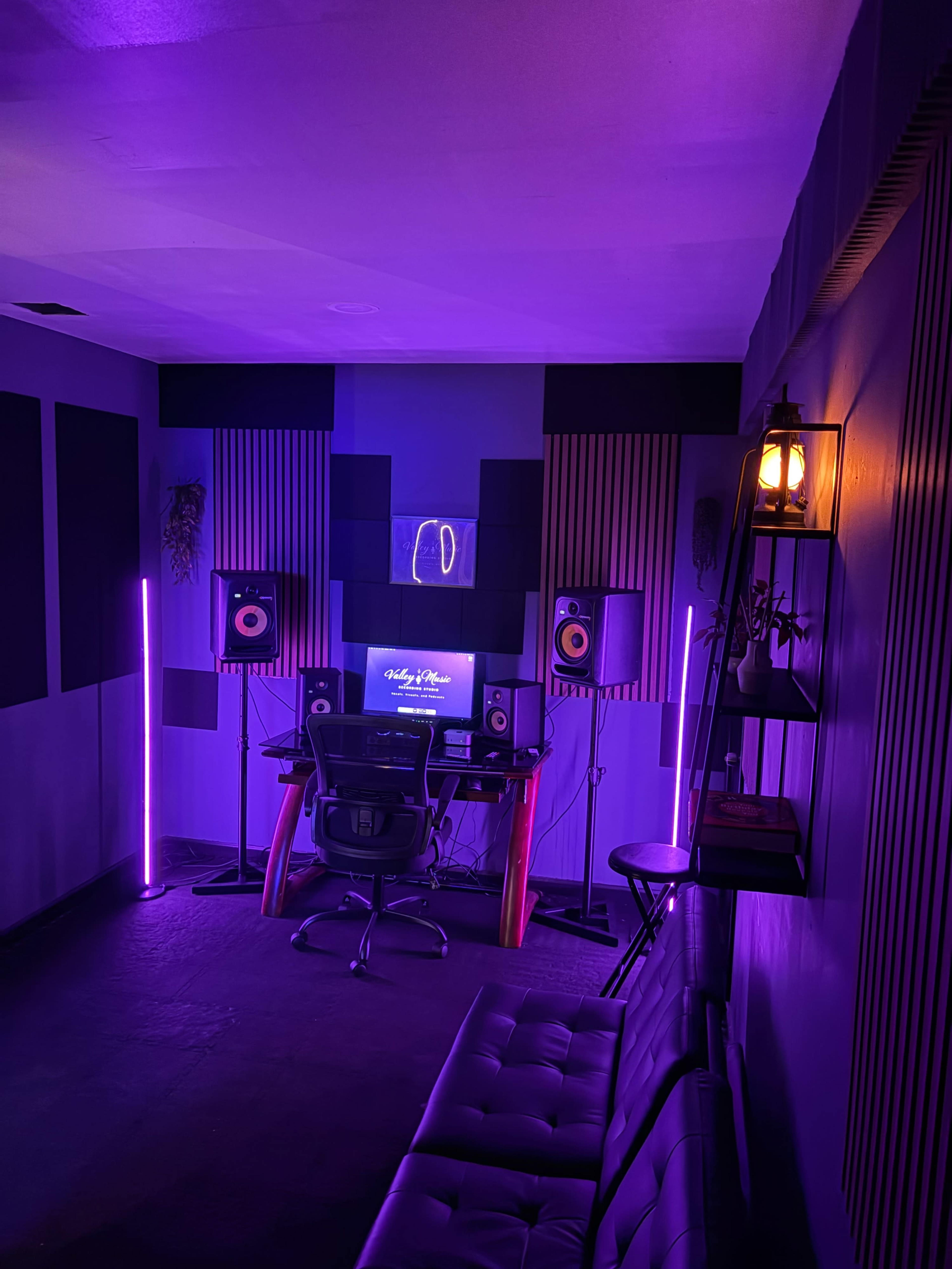 A small music studio features a desk with monitors, speakers, and ambient purple lighting, along with soundproofing panels and a seating area.
