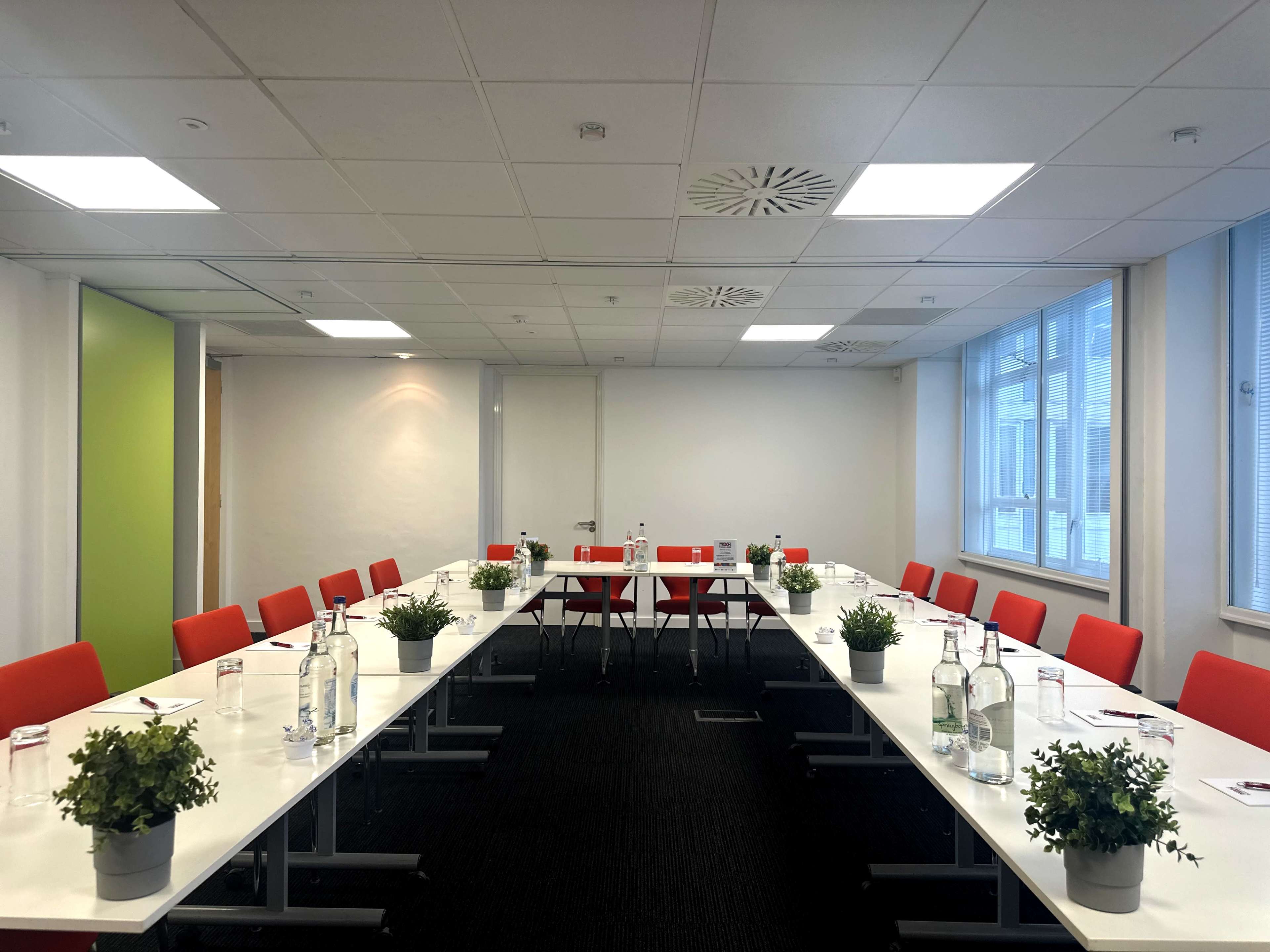 A modern conference room with a long table set for a meeting, featuring red chairs, water bottles, and small potted plants.