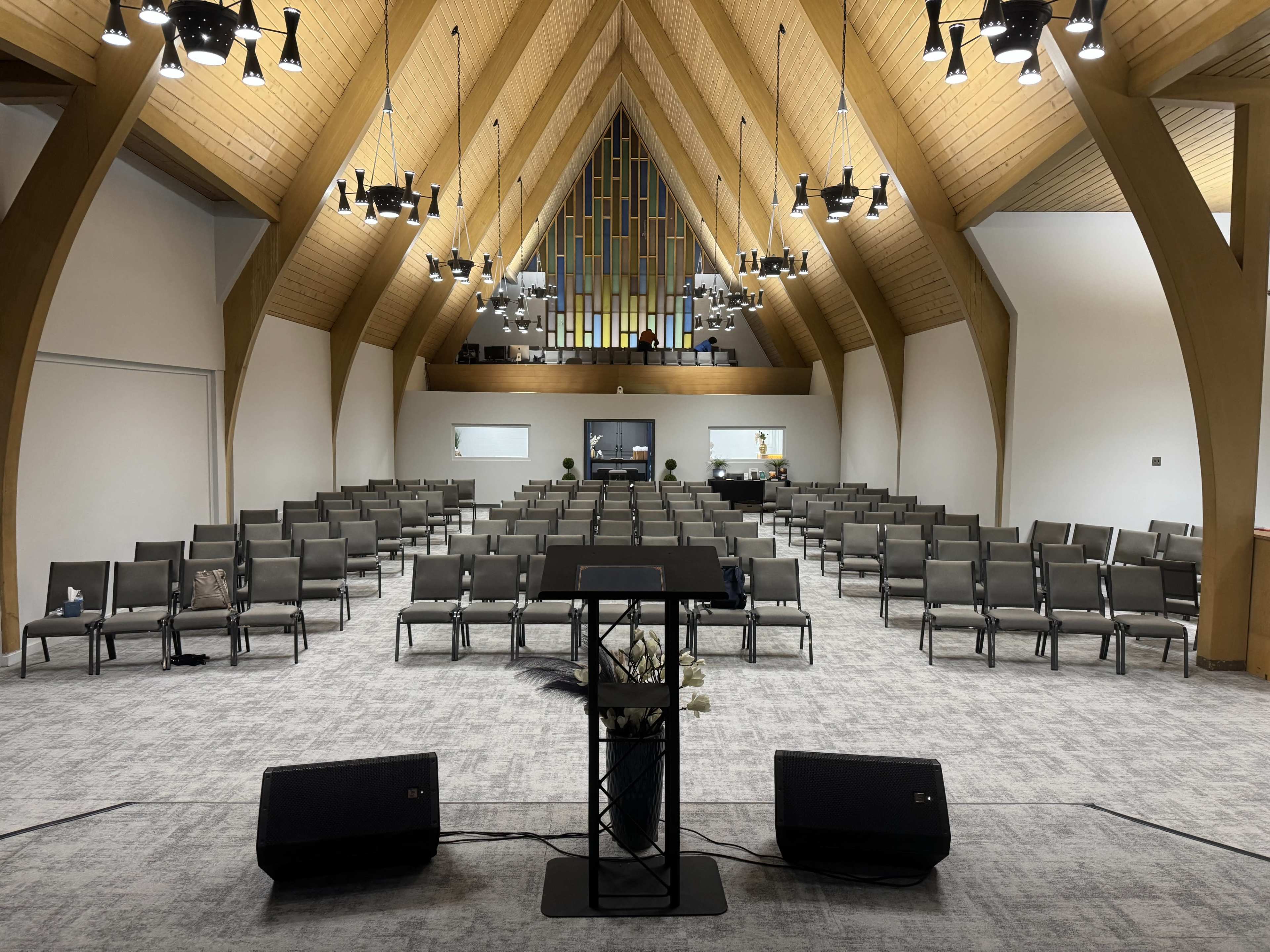 The image shows a modern church interior with a triangular wooden roof, rows of empty gray chairs, and a podium in the foreground.