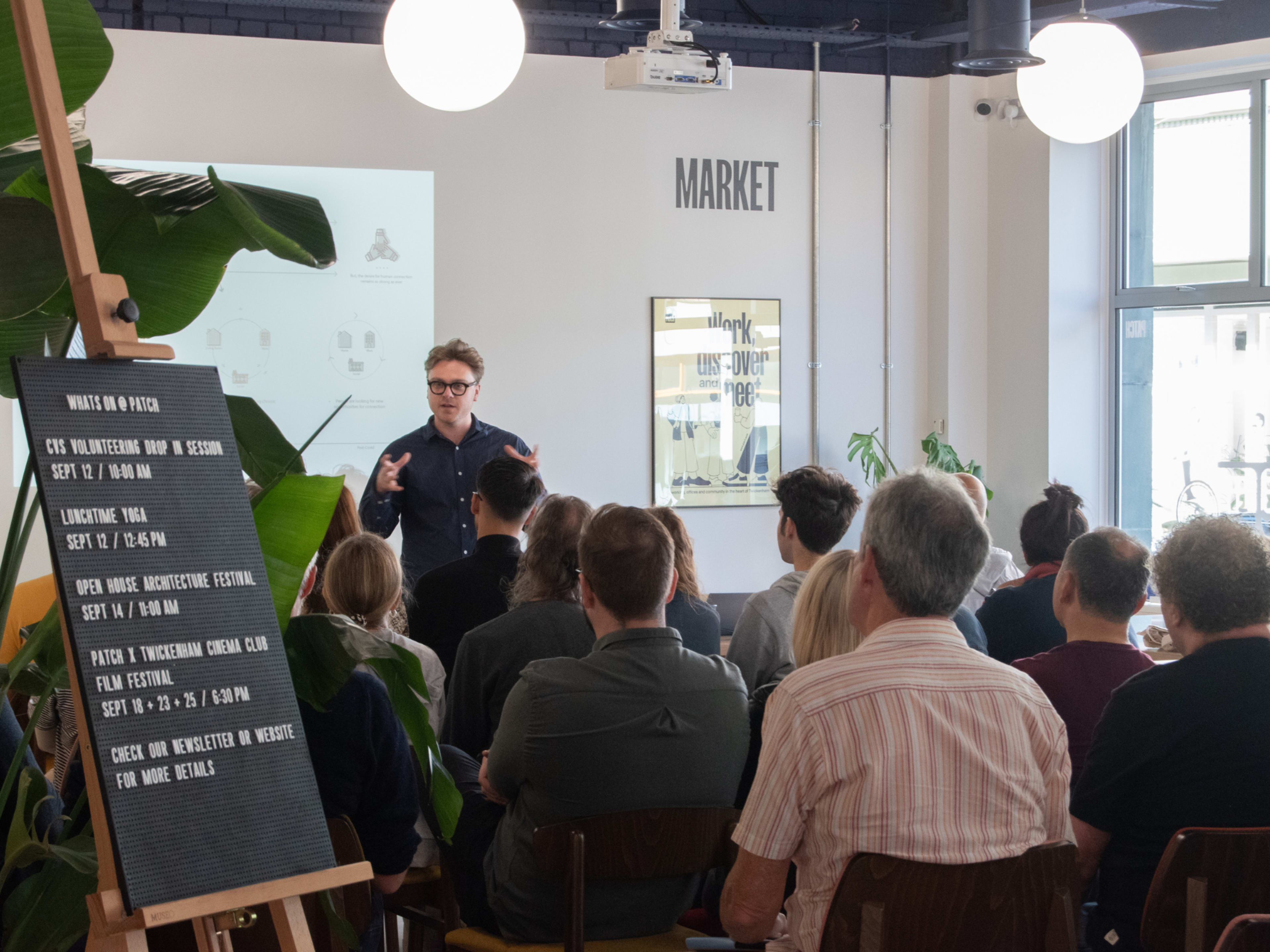 A speaker presents to an audience in a brightly lit room with plants, a marker board, and a sign that reads "MARKET."