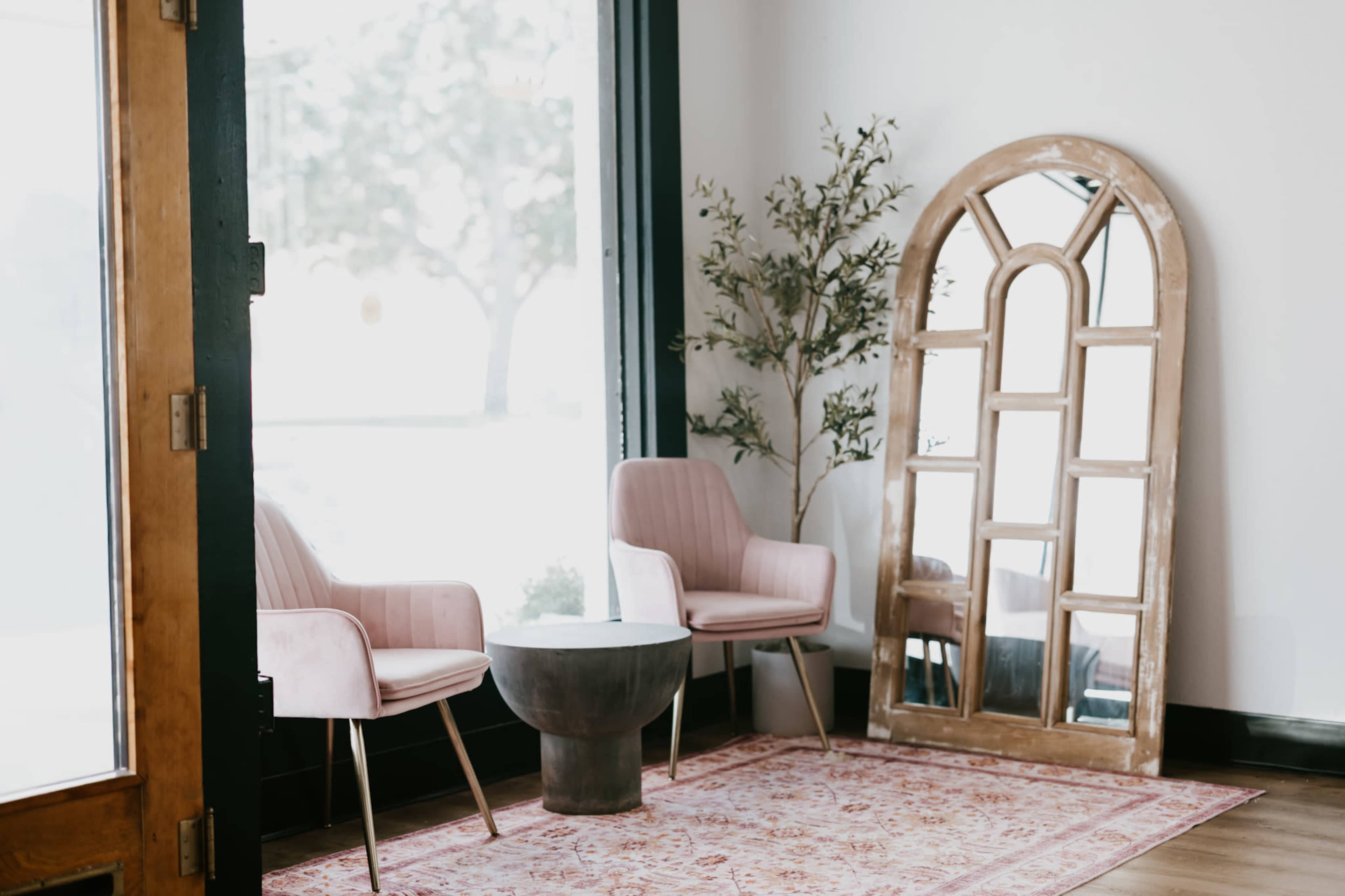 The image shows a modern interior space featuring two pink upholstered chairs, a round wooden coffee table, and a large arched mirror against a wall, all set on a patterned area rug.