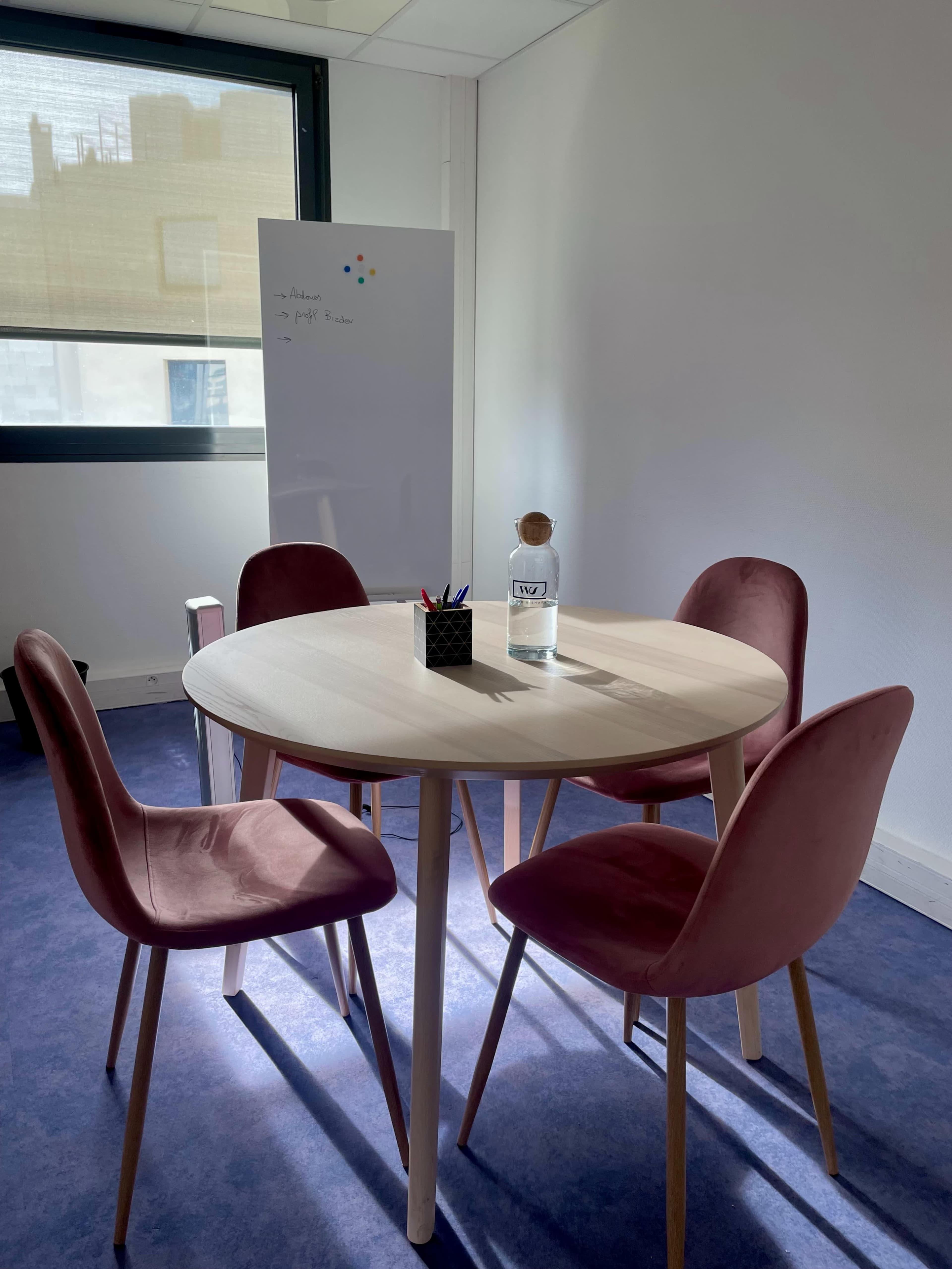 A round table with four pink chairs is set up in a bright office space next to a whiteboard.
