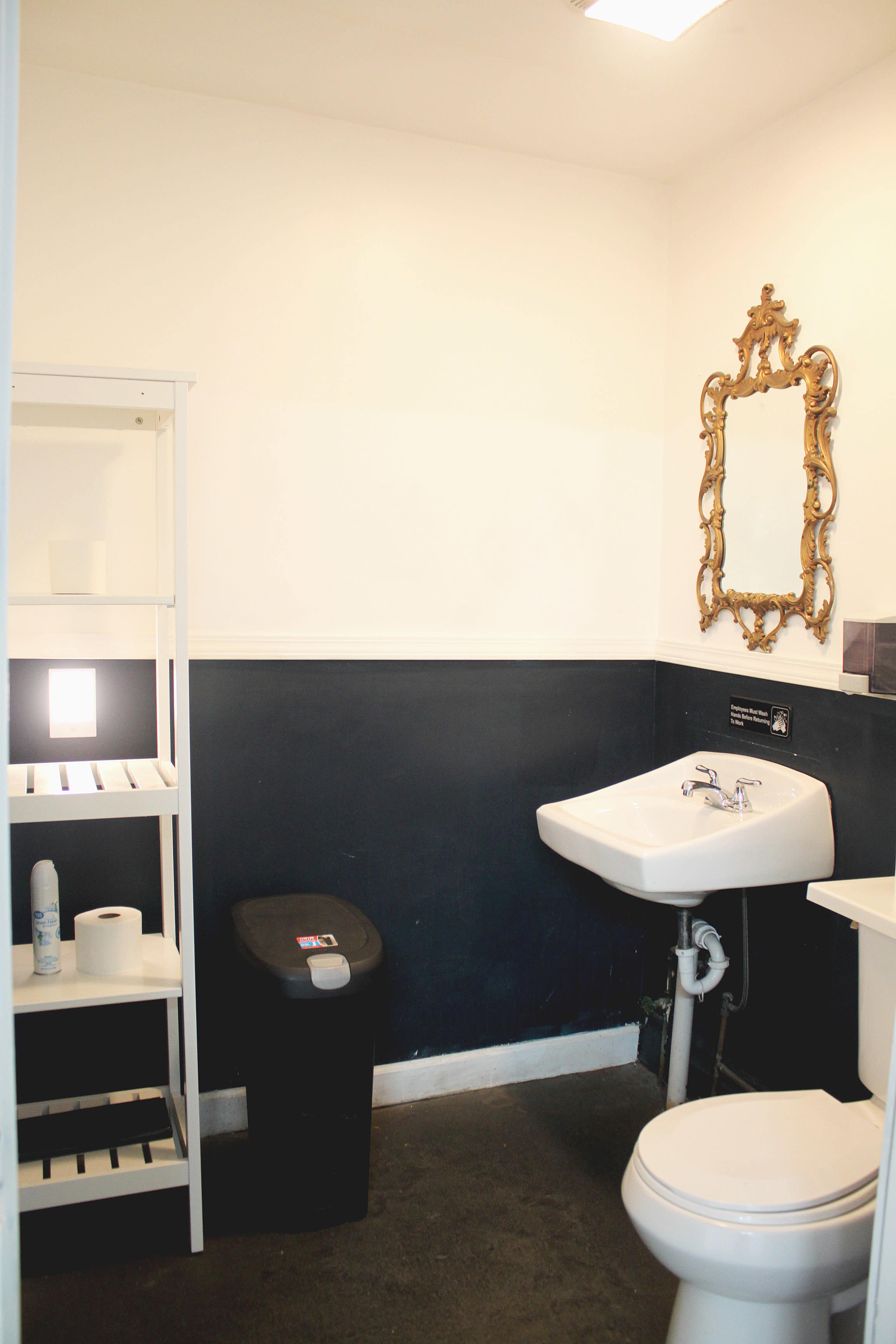 The image shows a small bathroom with a black and white color scheme, featuring a sink, toilet, mirror, and a shelf beside a trash bin.