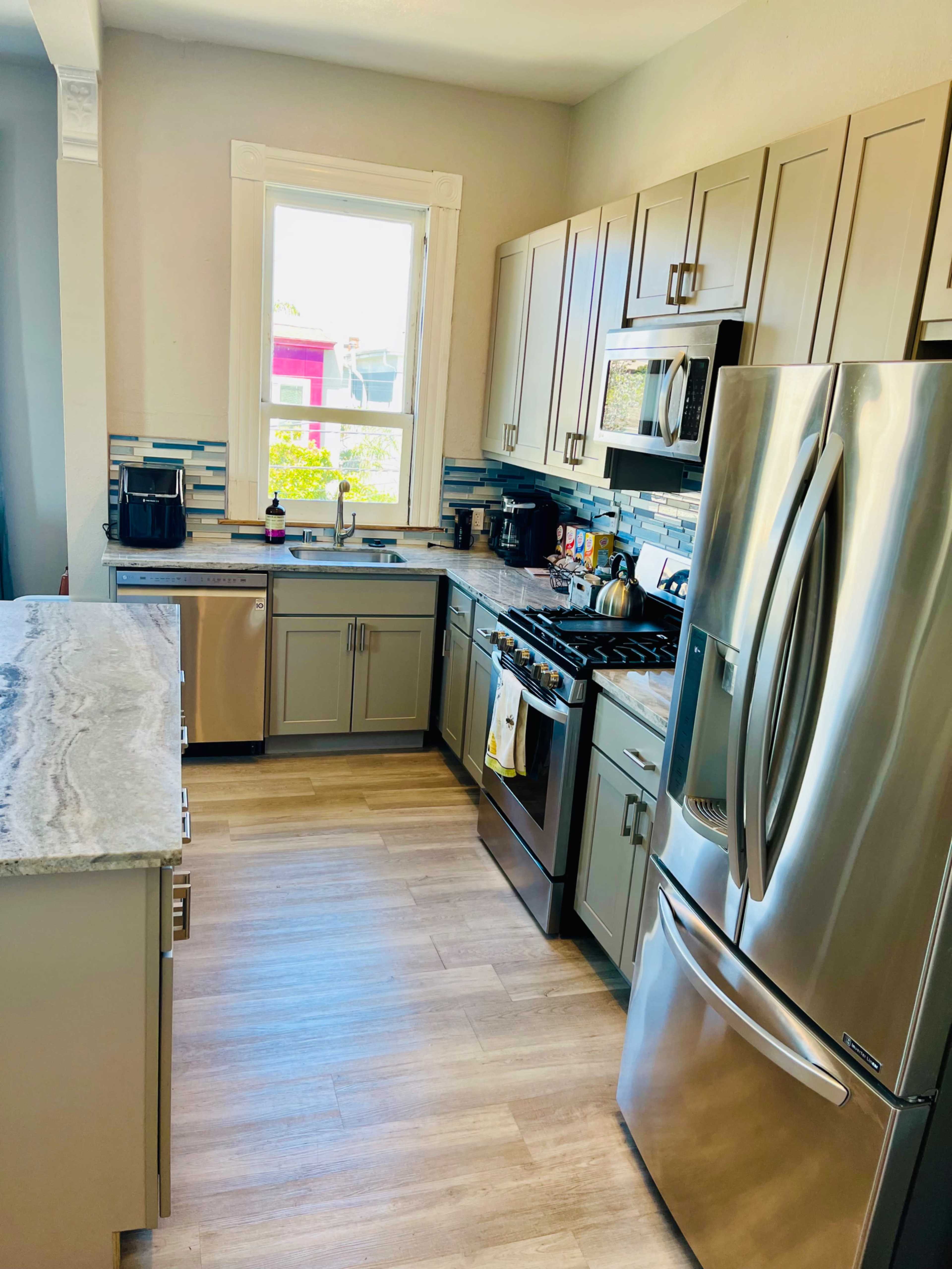 The image shows a modern kitchen featuring stainless steel appliances, light-colored cabinetry, and a window overlooking an outdoor view.