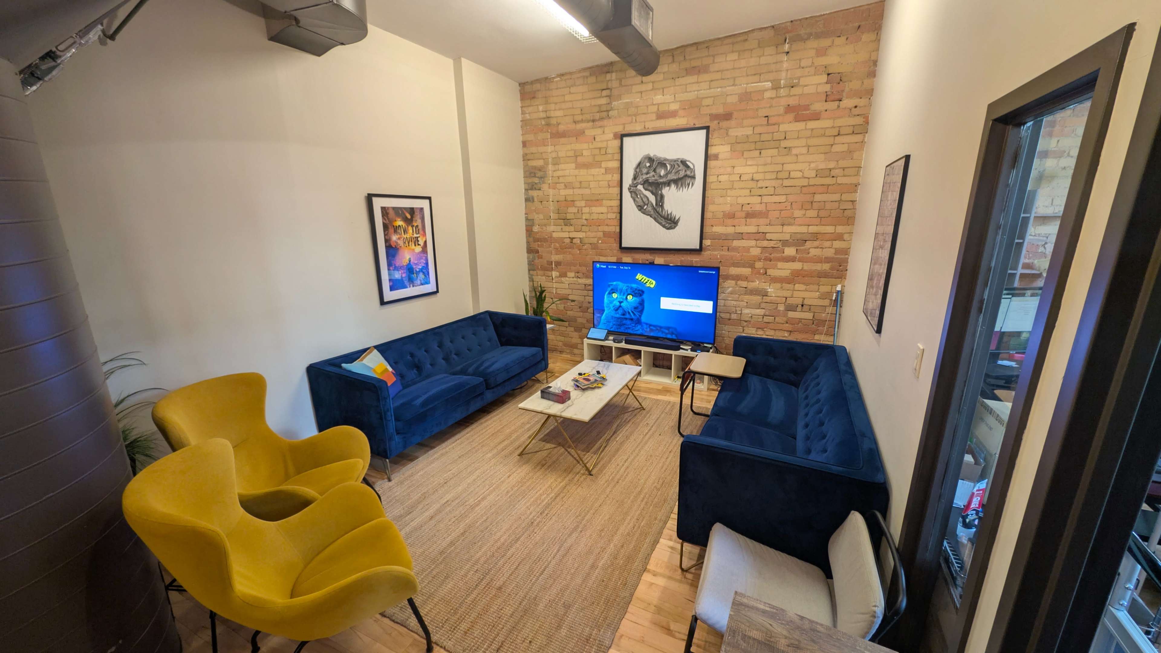 The image shows a modern lounge area with two blue sofas, a yellow armchair, a coffee table, and a large flat-screen TV, all set against a brick wall.