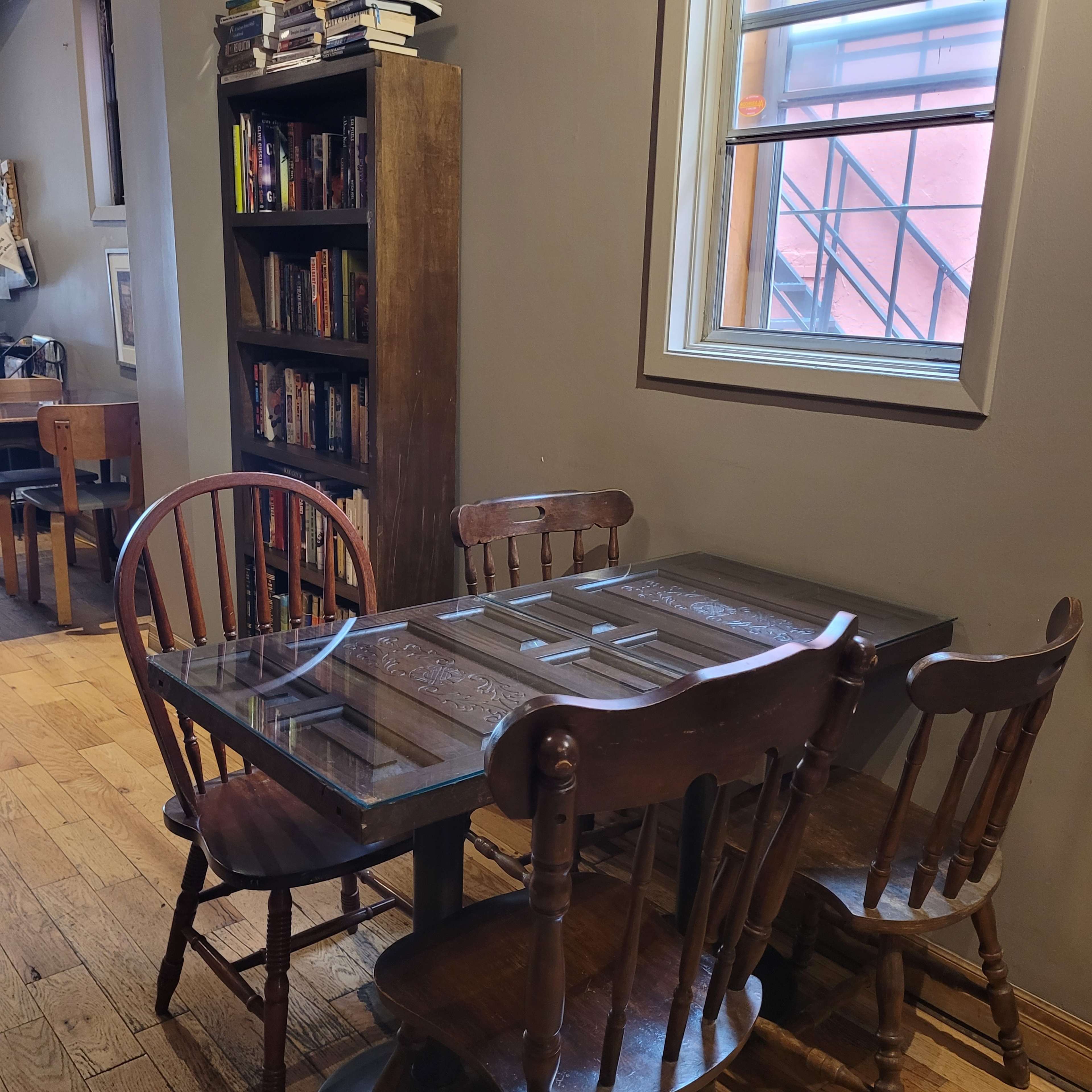 A small wooden table with four matching chairs beside a bookshelf filled with books and a window in a cozy room.