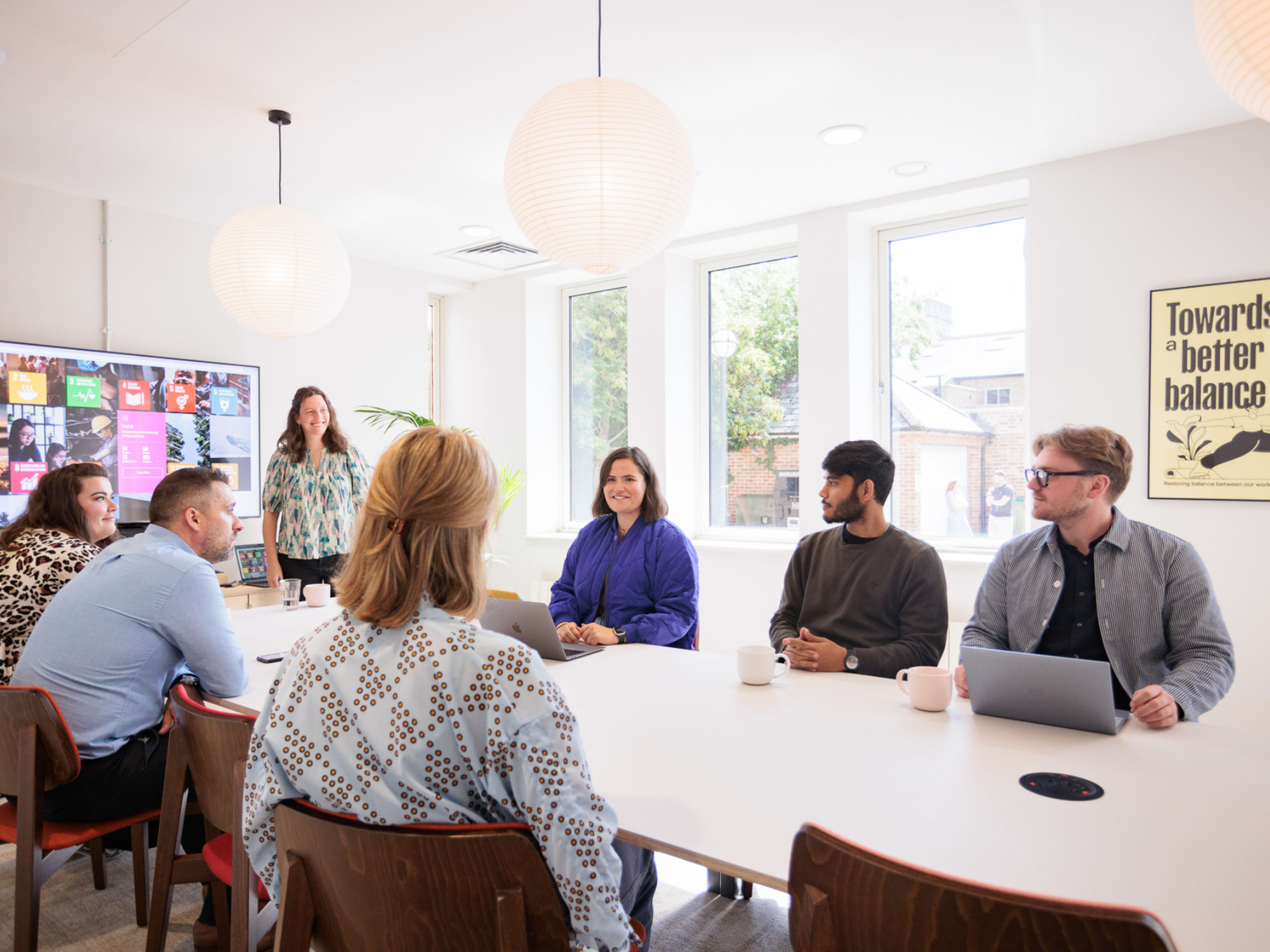A group of seven people sit around a conference table in a brightly lit meeting room, engaged in discussion, with a screen displaying images on the wall.