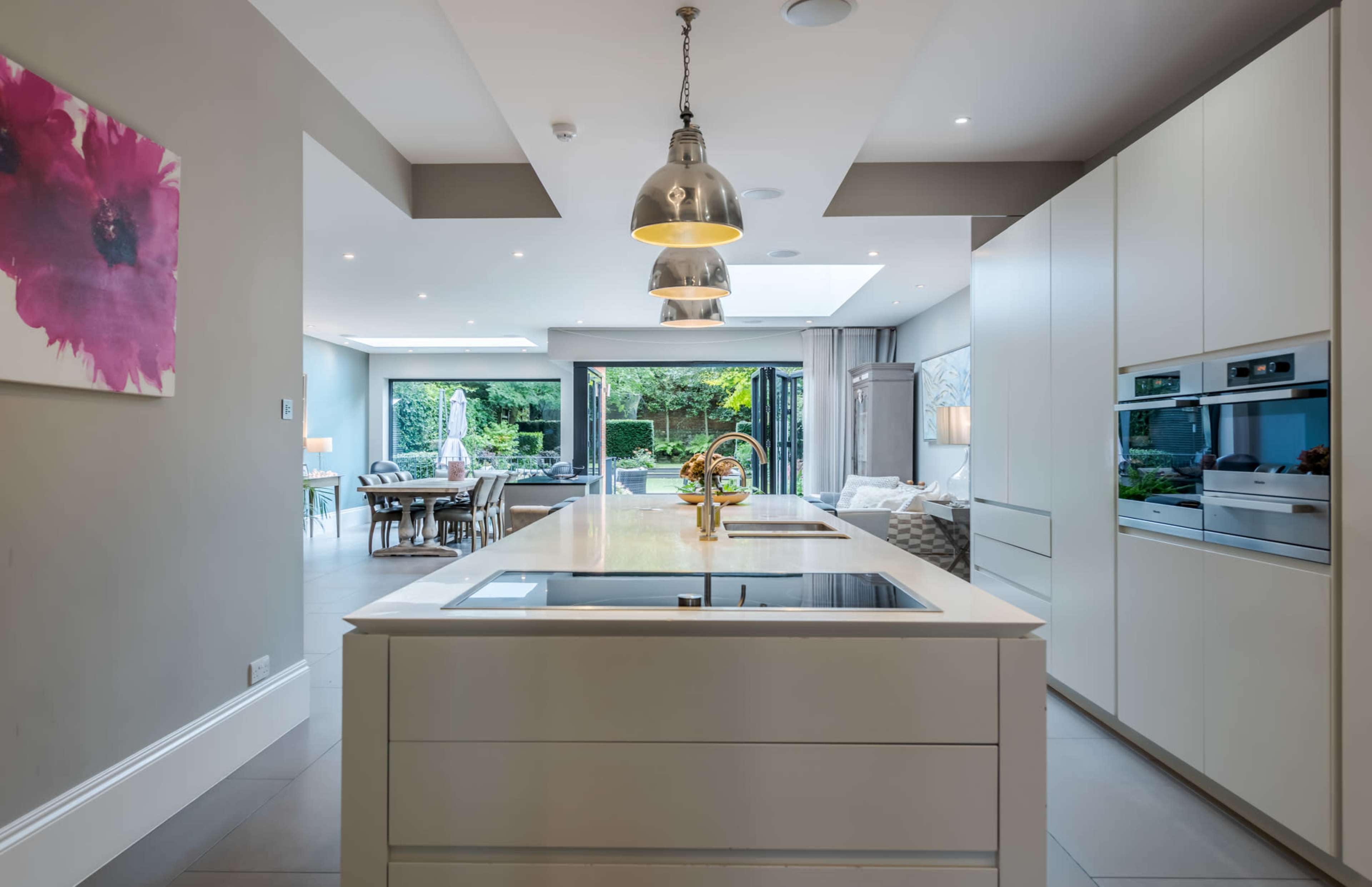 The image shows a modern kitchen with an island, built-in appliances, and large windows that overlook a garden area.