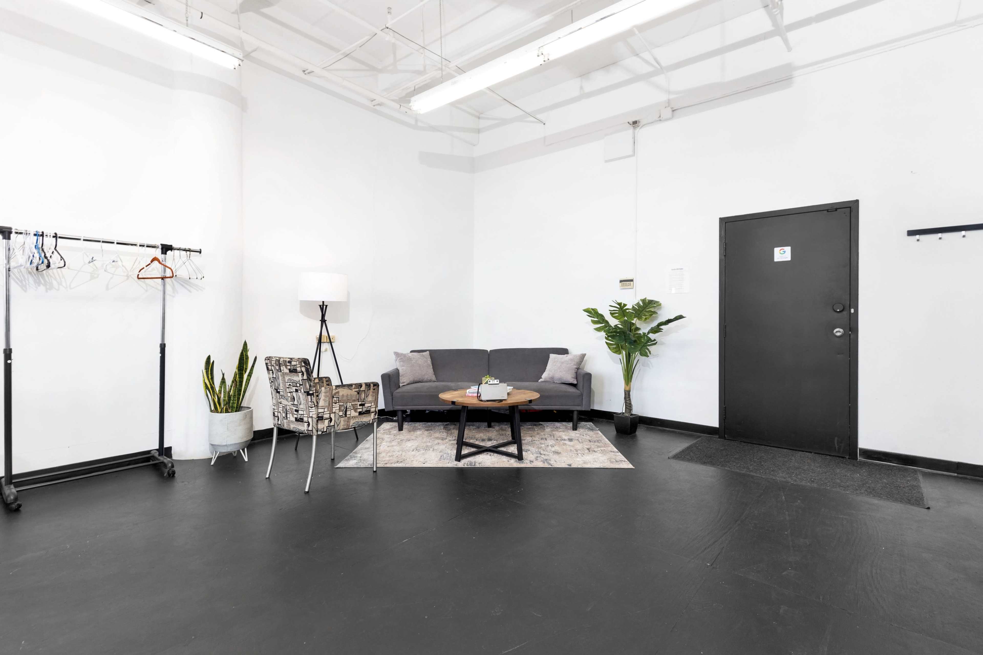 A modern, minimalistic room with a gray couch, a black door, a small table, a floor lamp, a plant, and a clothing rack against the white walls.