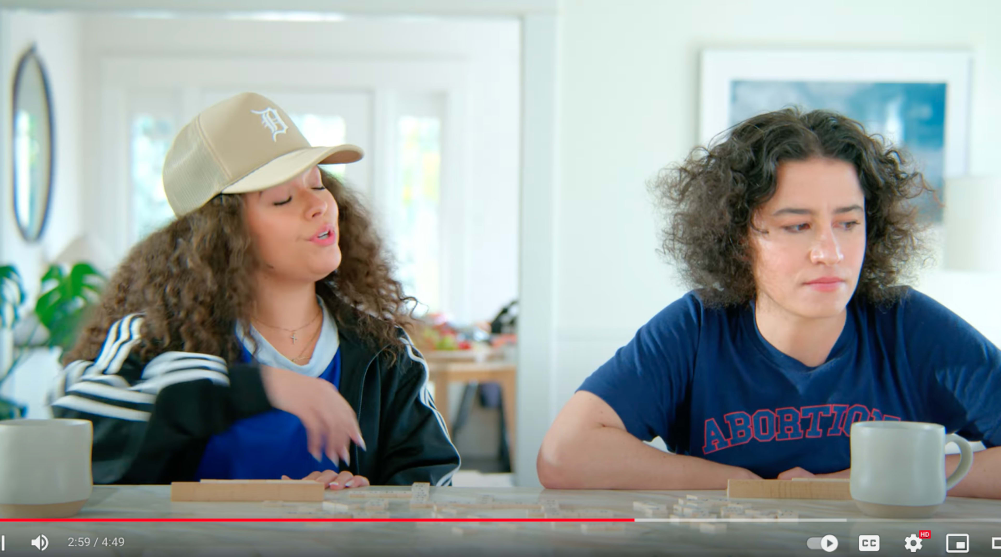 Two young women sit at a table, engaged in conversation, with a bright, modern interior in the background.