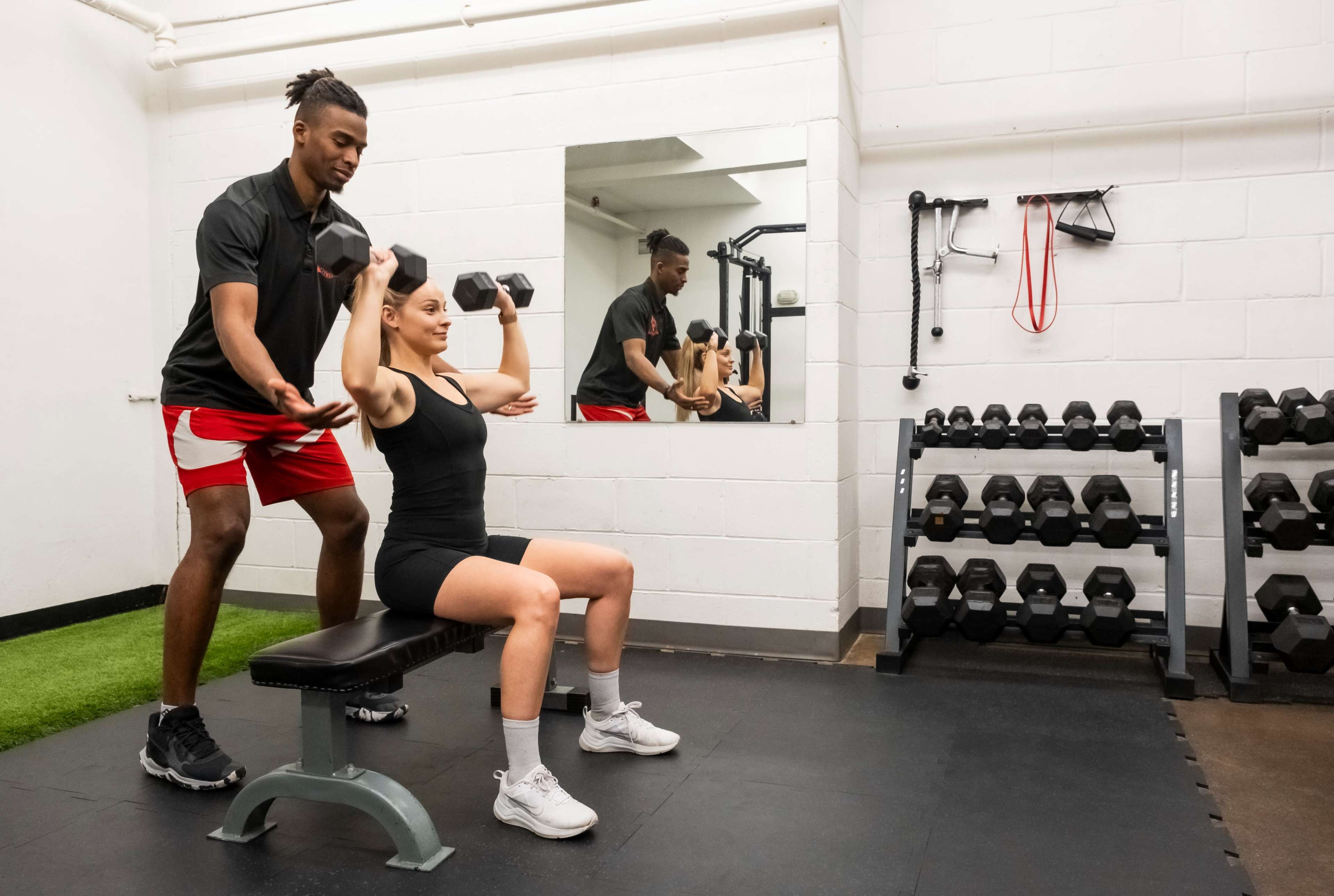 A fitness trainer assists a woman with her overhead dumbbell press on a bench in a gym equipped with weights.