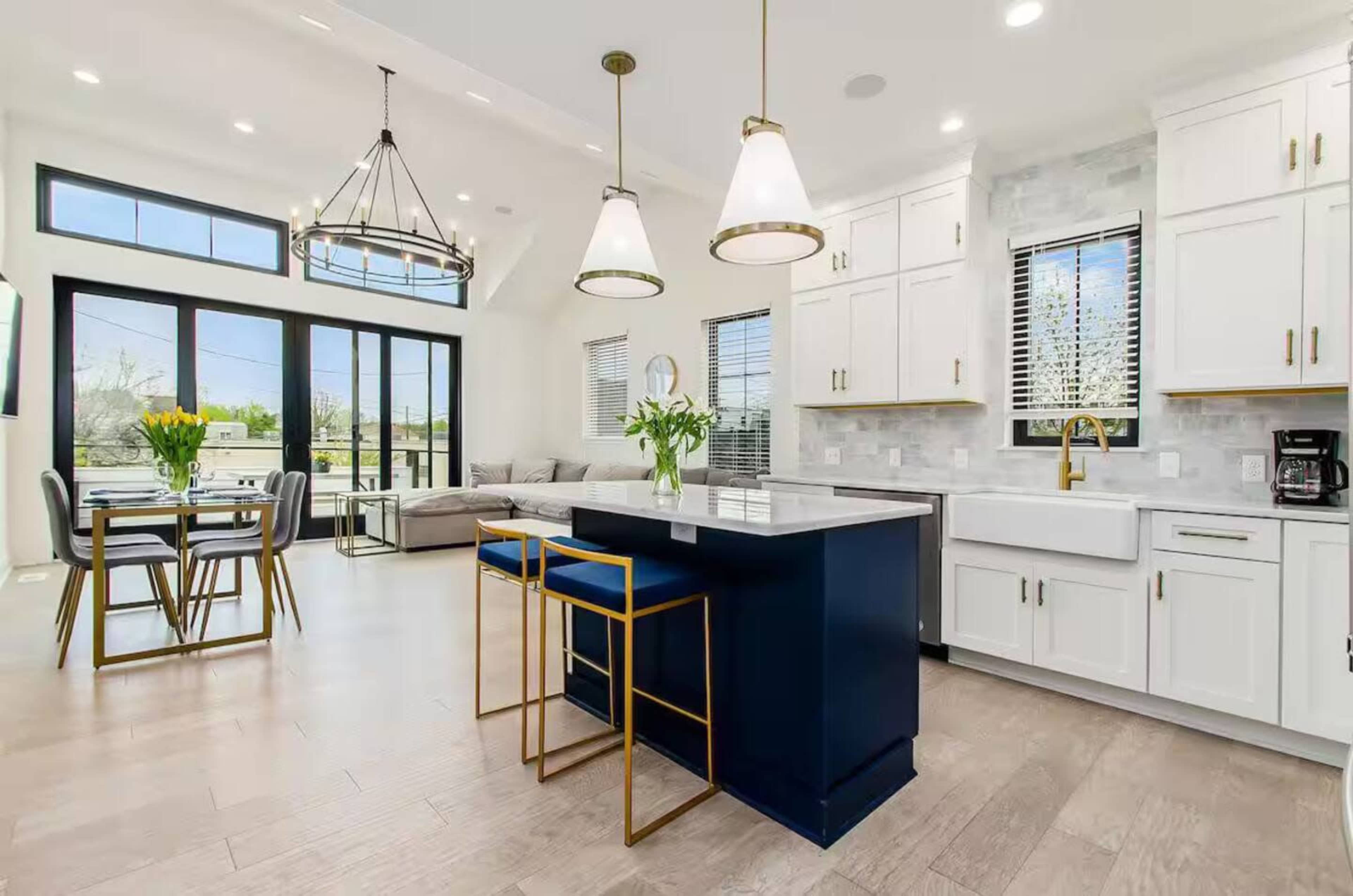 The image shows a modern kitchen and dining area featuring white cabinetry, a blue kitchen island with bar stools, a dining table set for four, and large windows providing natural light.