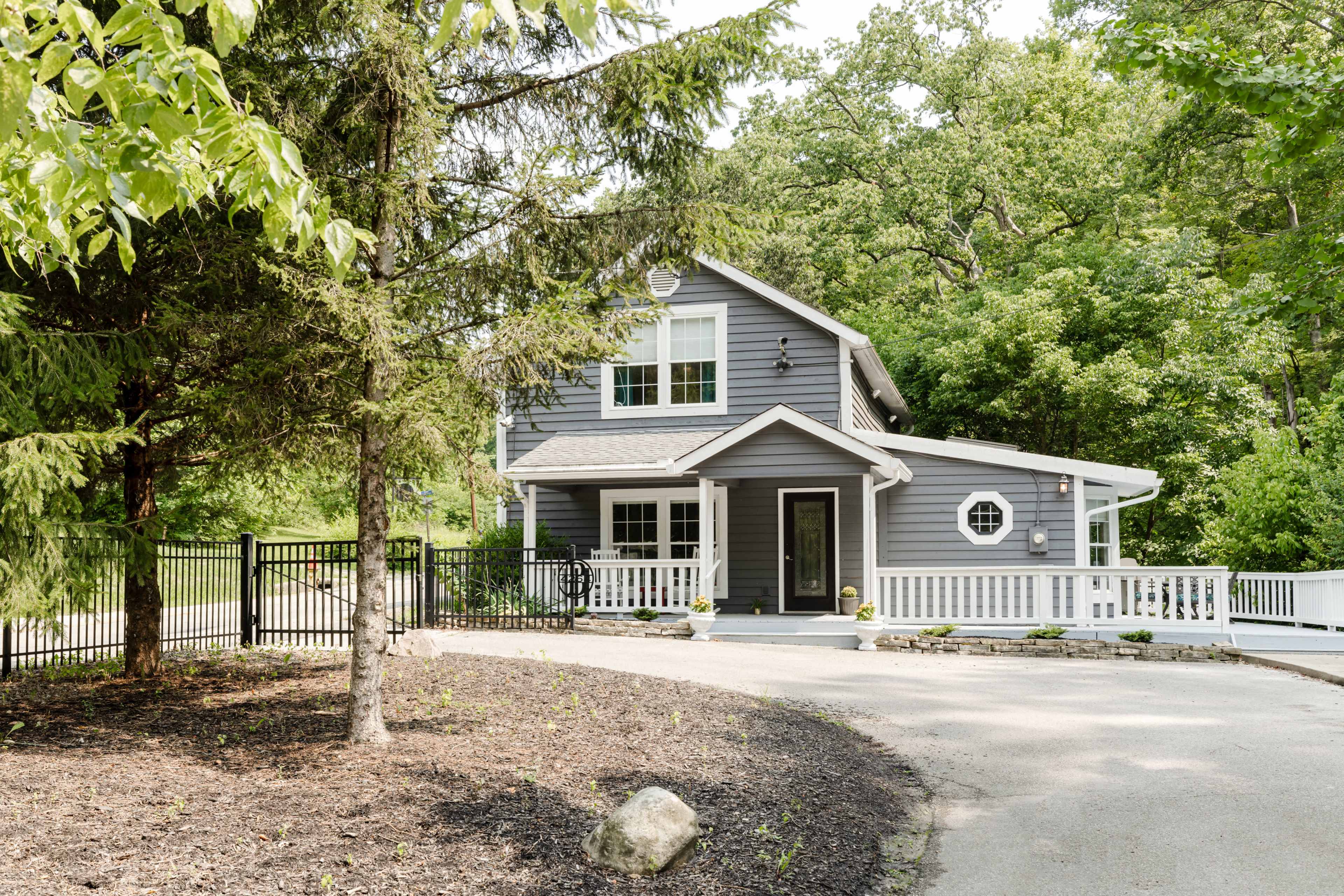 A two-story gray house with white trim is surrounded by greenery and features a curved driveway and a fenced front yard.
