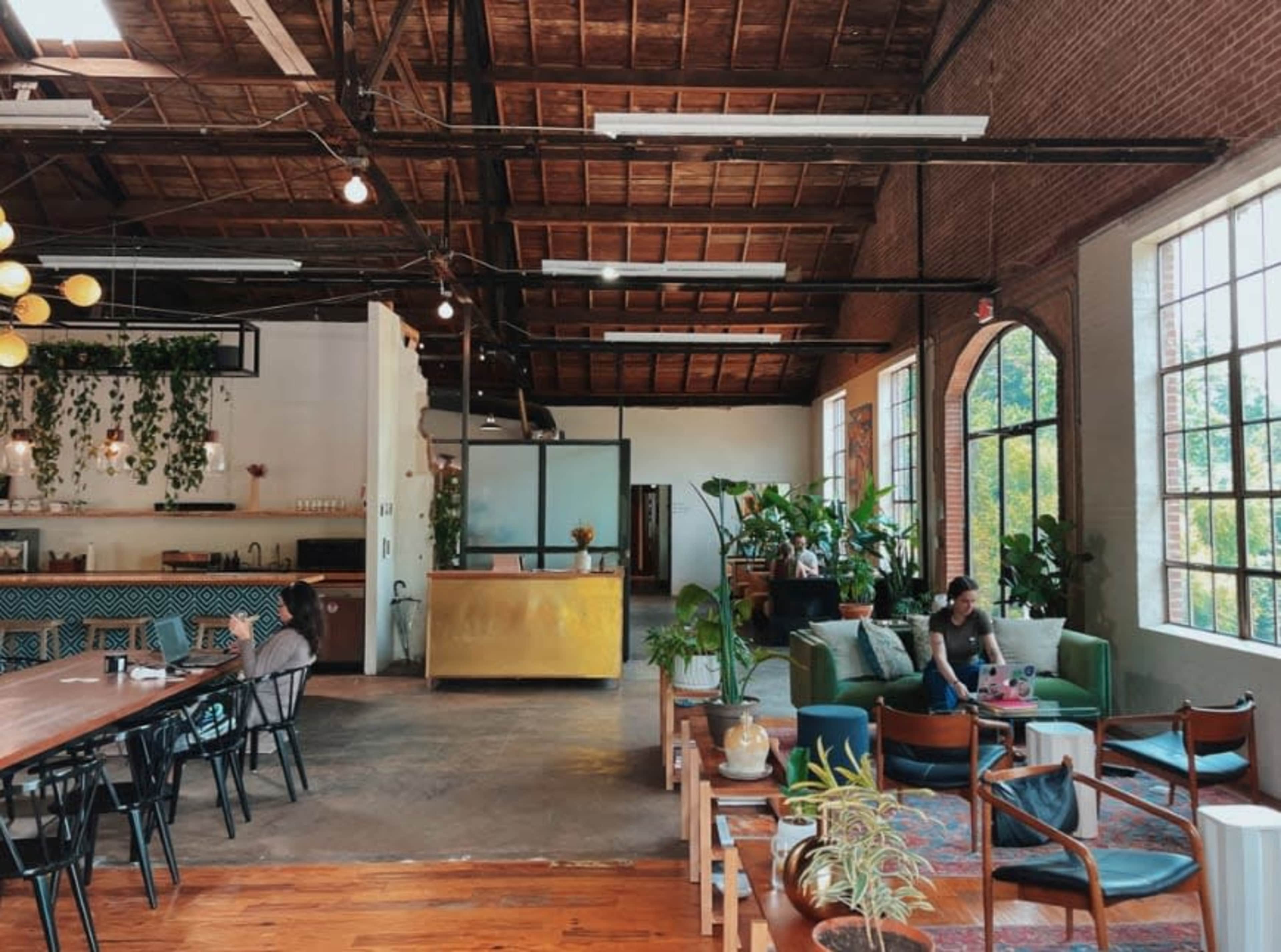 The image shows an open, spacious café with high ceilings, large windows, and various seating areas filled with plants and greenery.