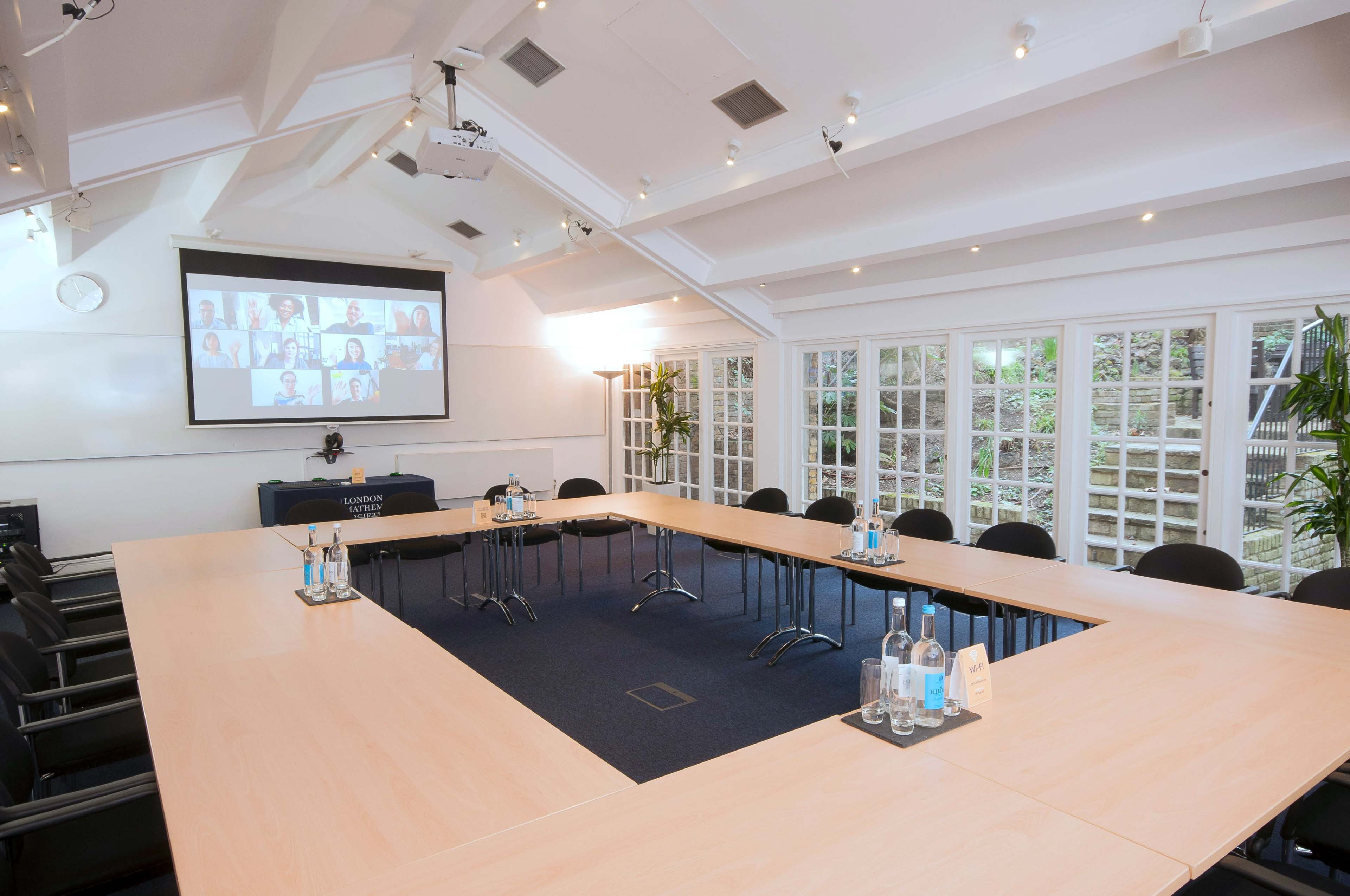 A conference room is set up with a large rectangular table, chairs arranged around it, and a screen displaying a virtual meeting in the background.