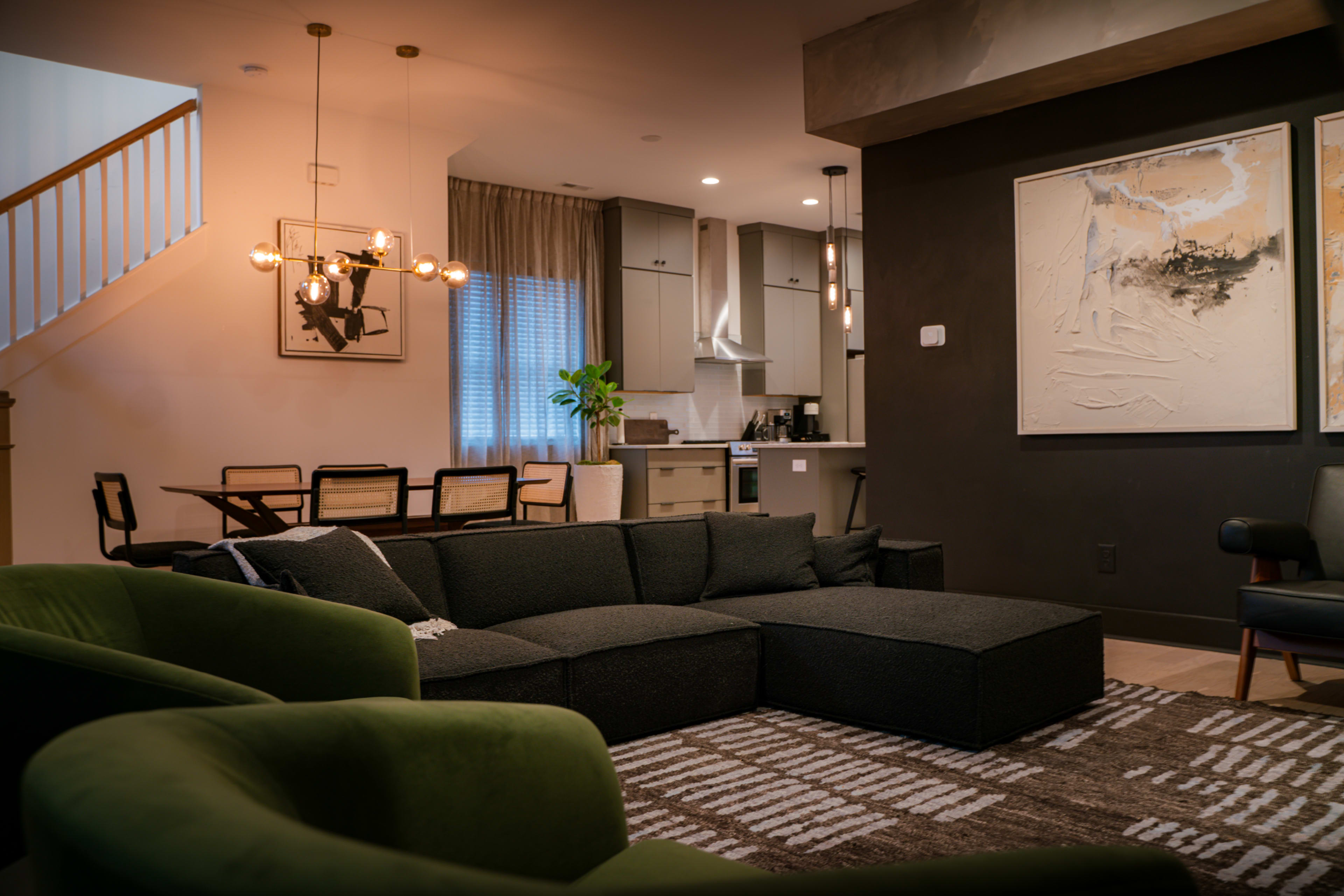 A modern living room with a dark sectional sofa, two green armchairs, a patterned rug, and a dining area in the background featuring a large table and kitchen.