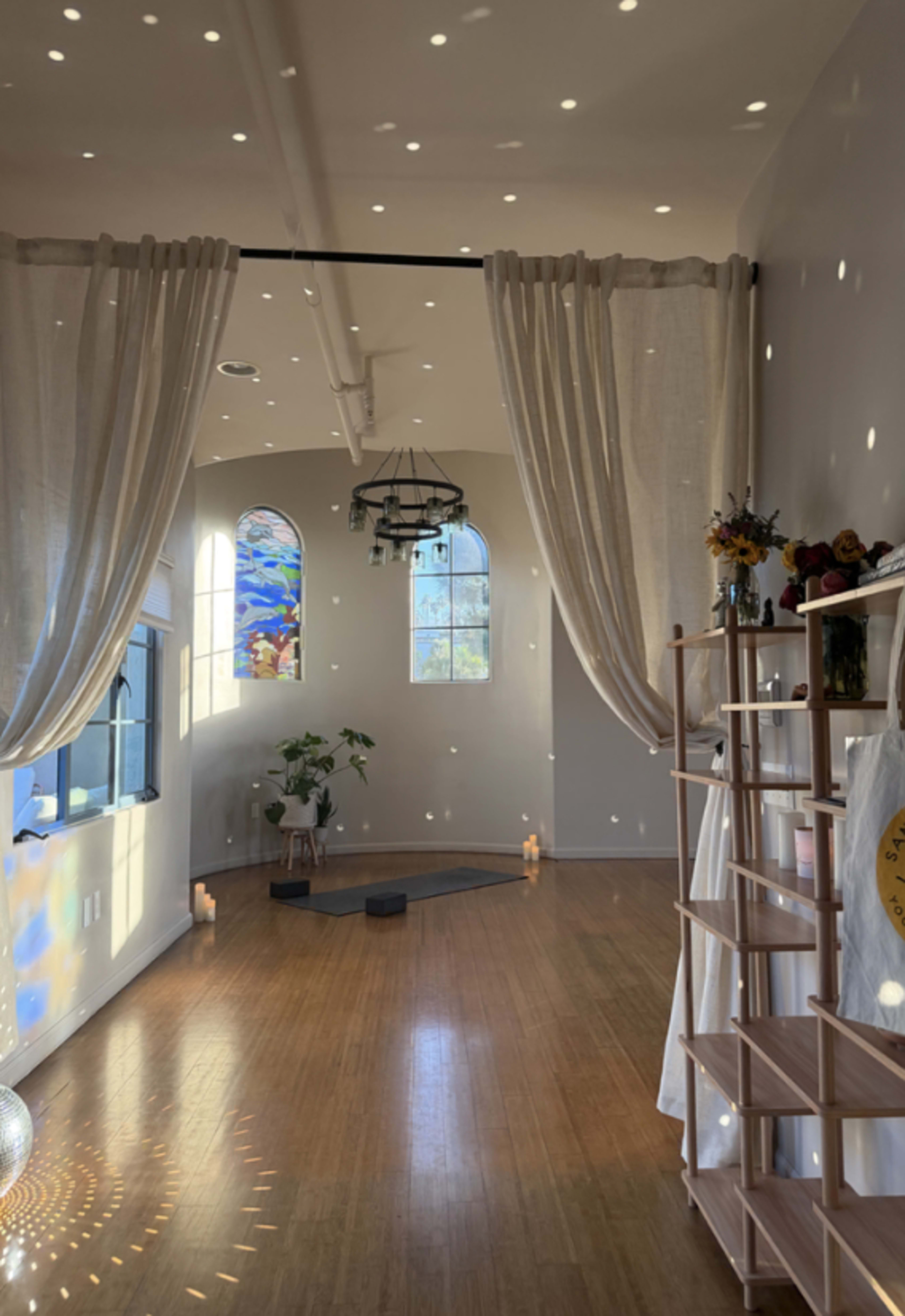 The image shows an inviting, airy space with large windows, soft curtains, and a wooden floor, illuminated by light reflections on the walls and floor.
