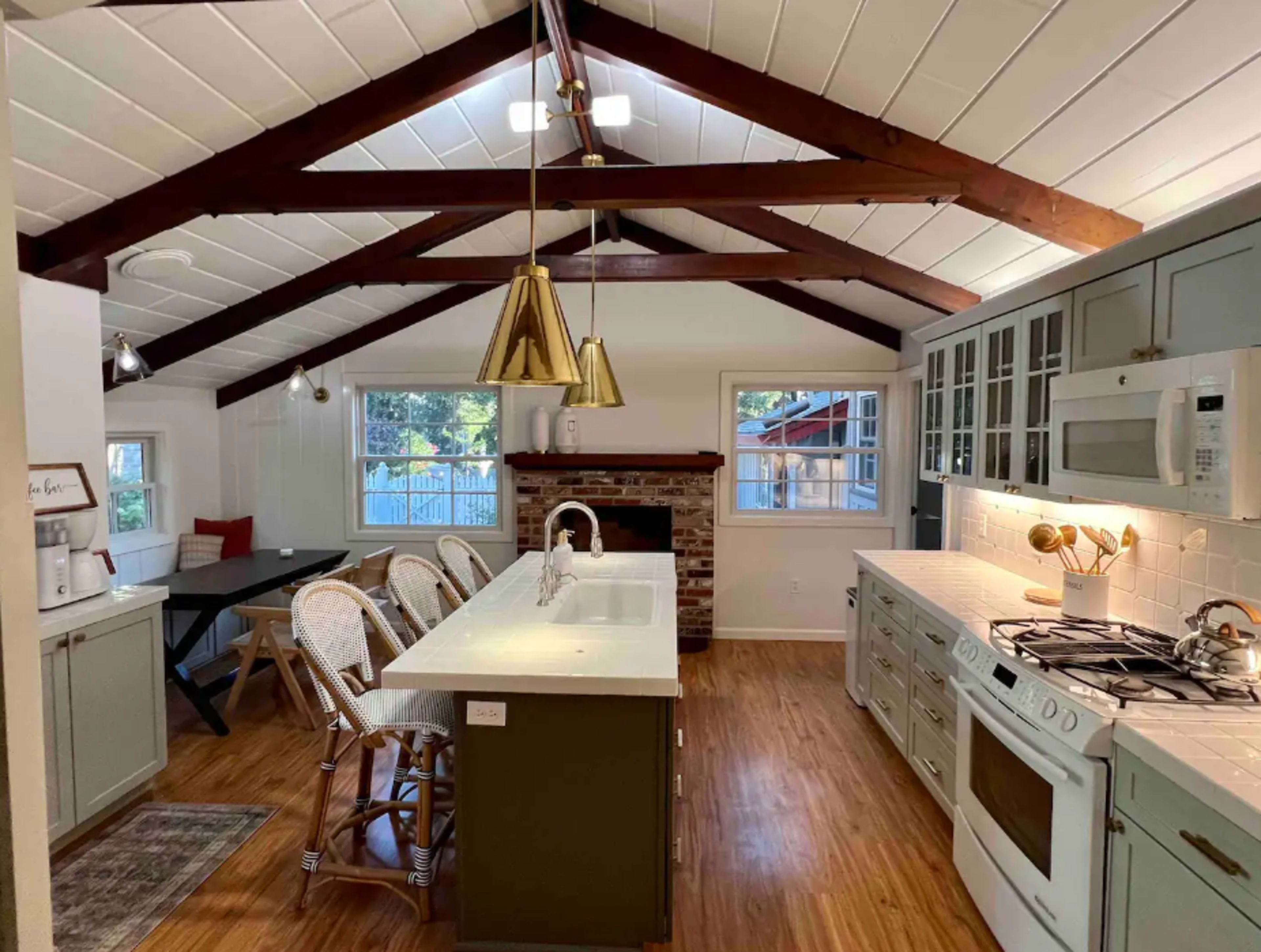 The kitchen features a high ceiling with exposed beams, a central island with seating, and modern appliances, alongside a brick fireplace.