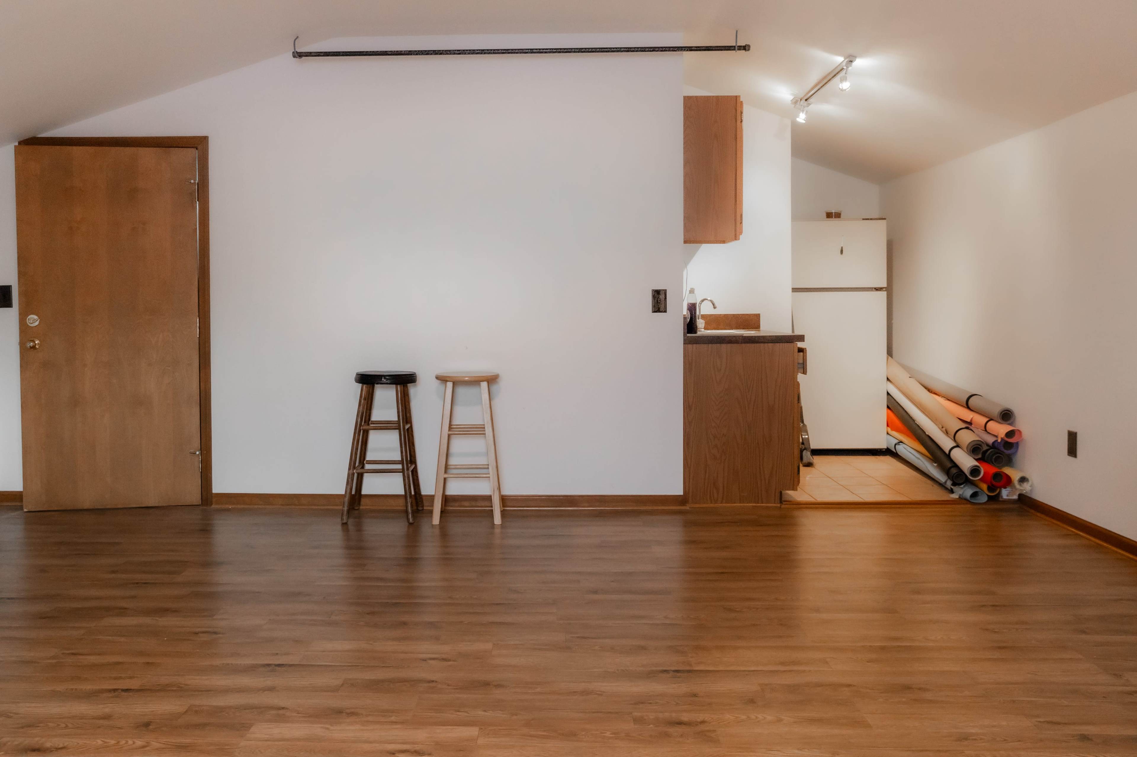 A sparsely furnished room with two stools and a kitchenette in the background, along with rolls of material resting against the wall.