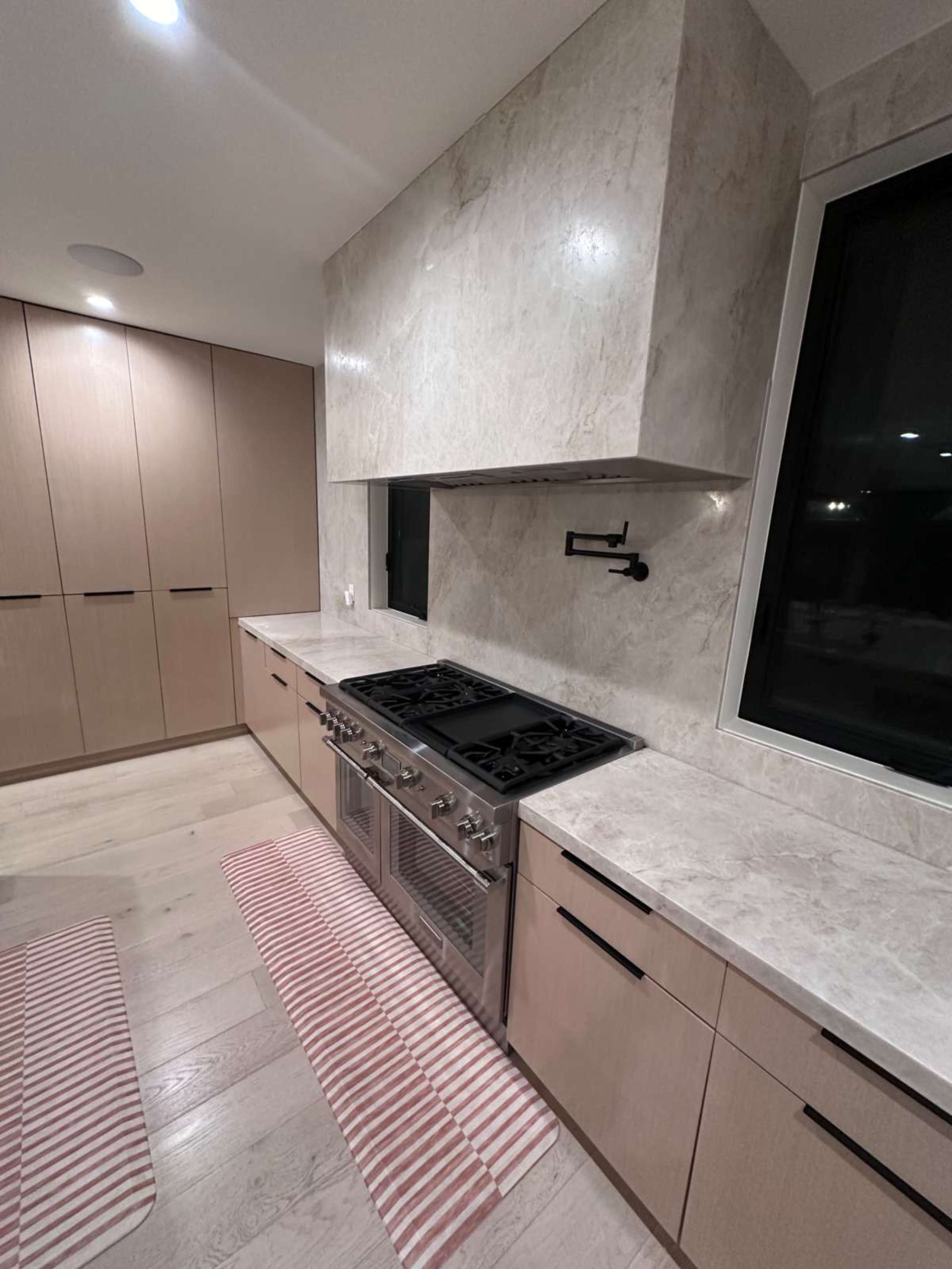 The kitchen features a marble backsplash, a large stainless steel gas stove, and light wood cabinetry with pink striped rugs on the floor.