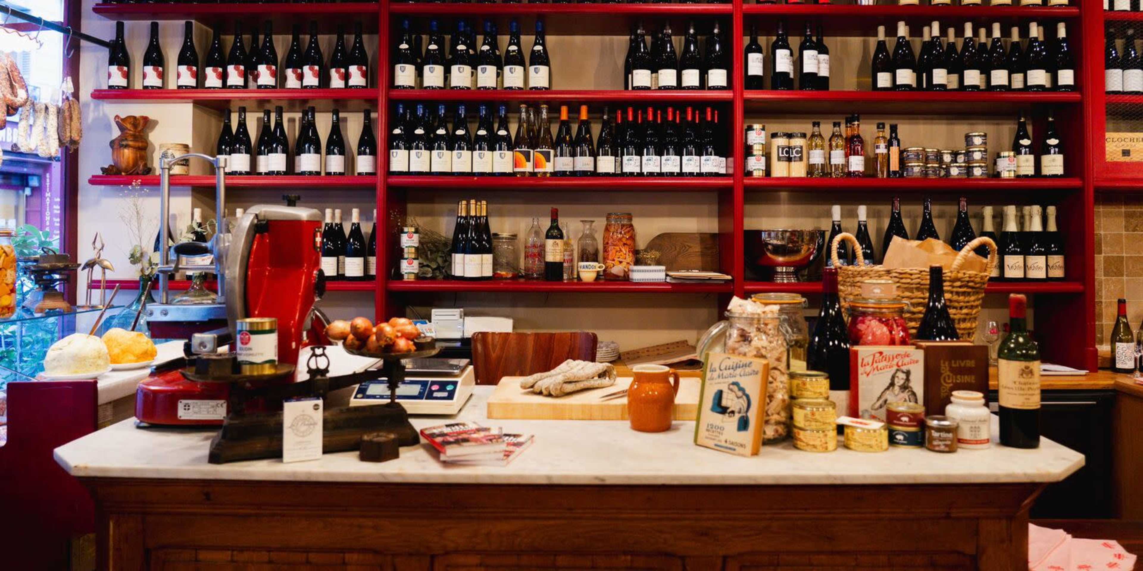 The image shows a rustic shop counter with various bottles of wine on shelves above, along with jars, baskets, and kitchen tools arranged on the countertop.
