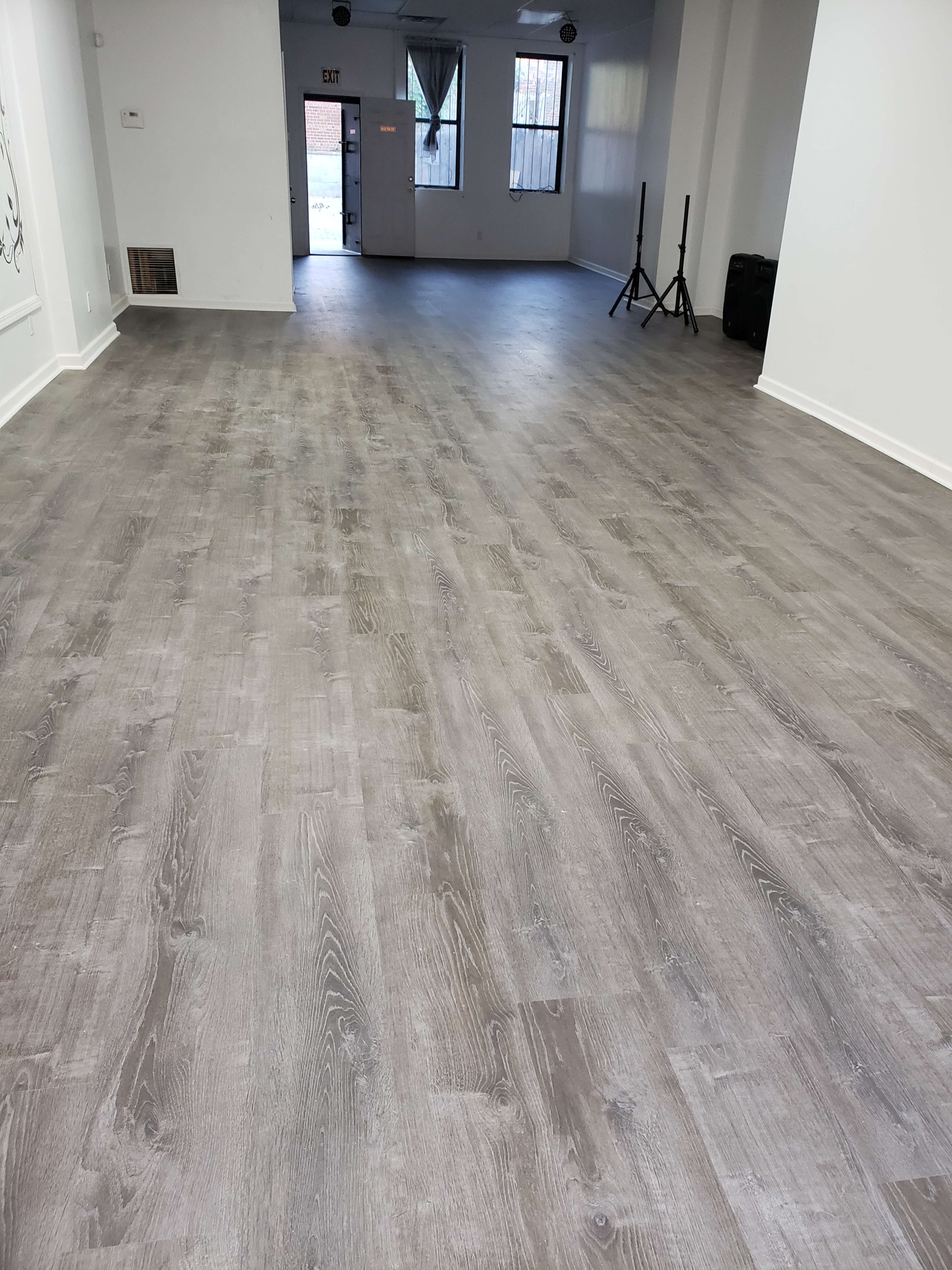 A spacious room with gray wooden floors, white walls, and large windows letting in natural light.