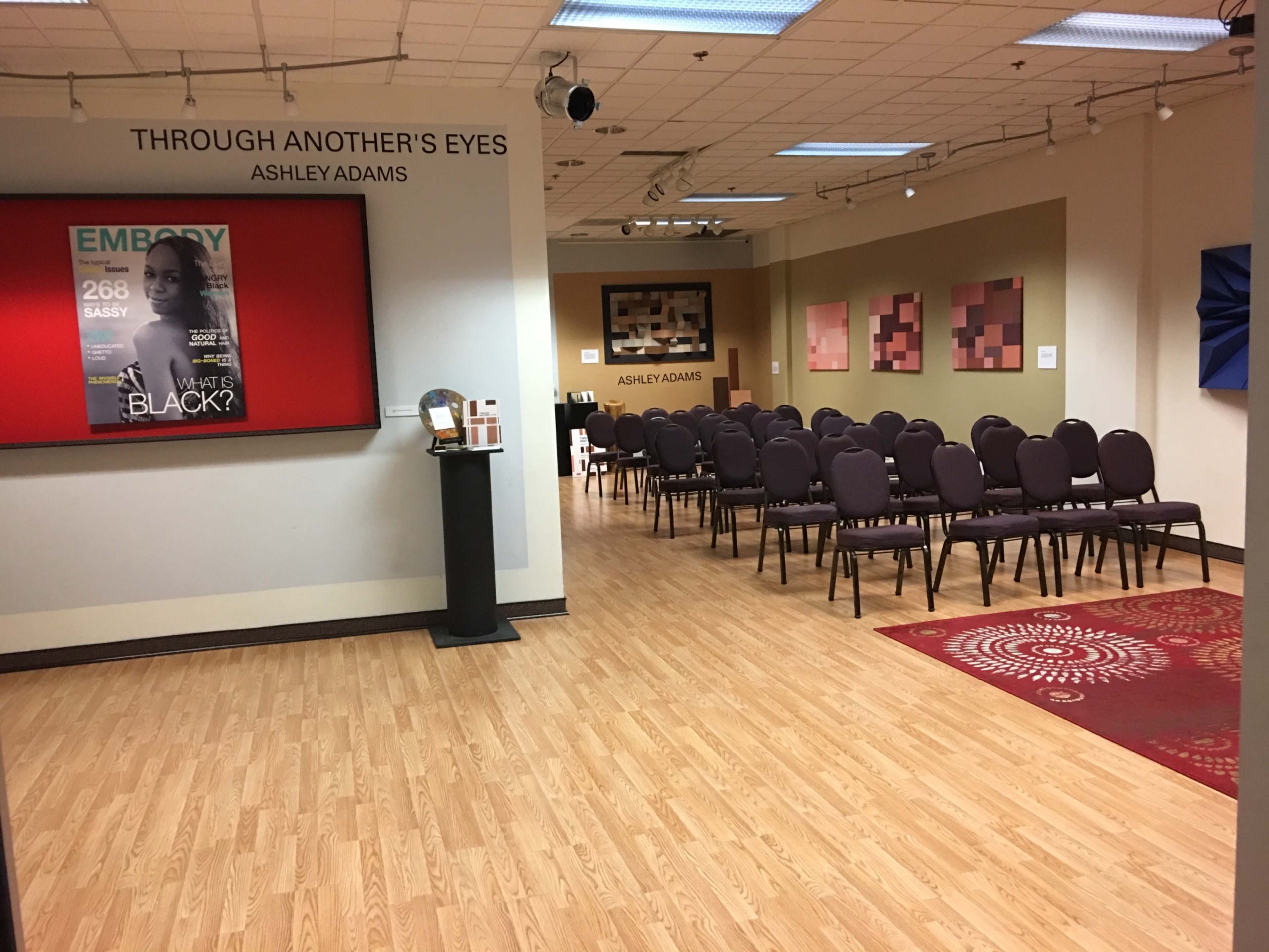 A hallway leading to an exhibition space with a display titled "Through Another's Eyes" and a set of empty chairs arranged for an event.