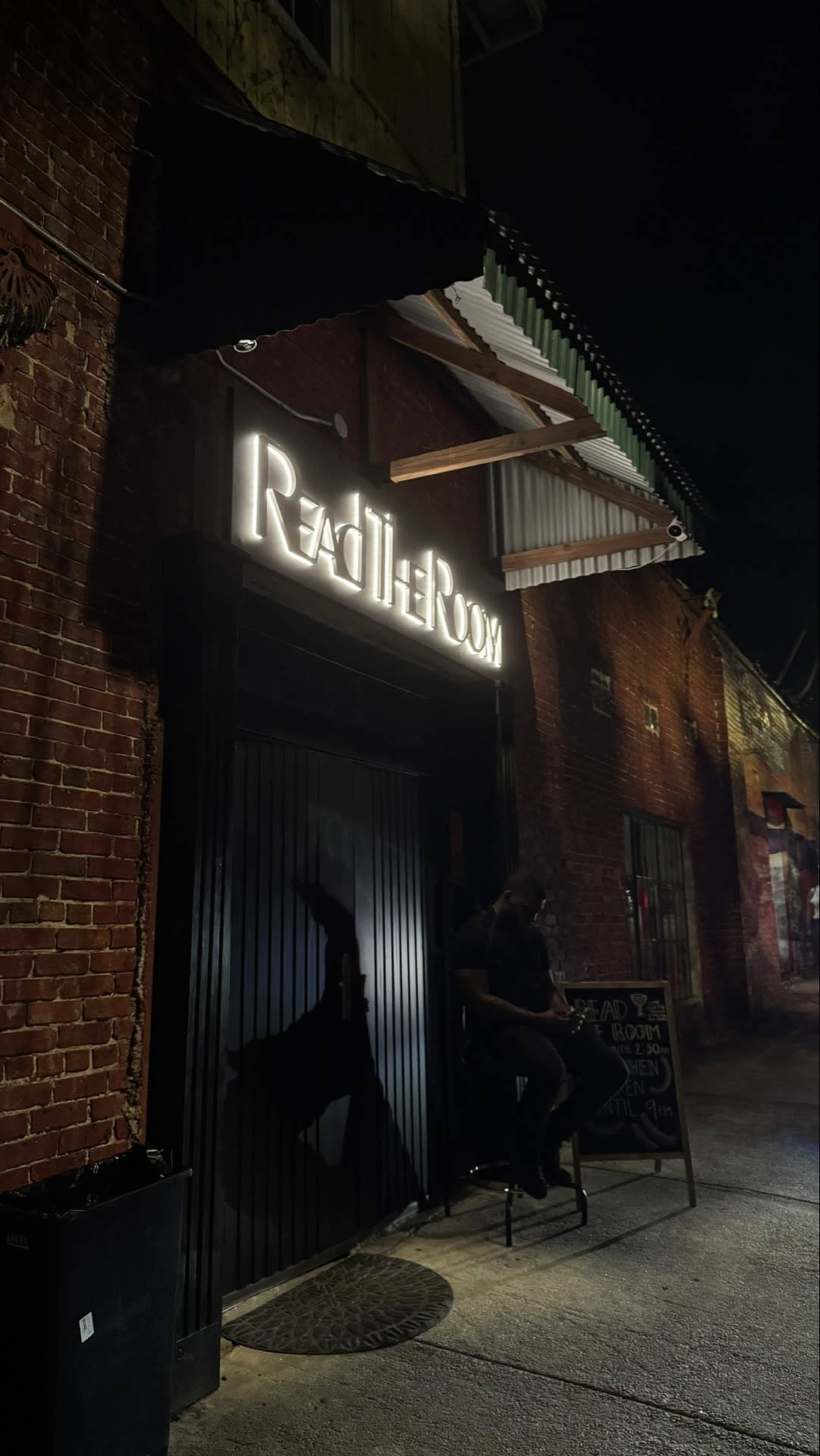 A person sits on a stool in front of a dimly lit entrance to a venue named "Reach The Room," with a brick wall backdrop and a sidewalk sign nearby.