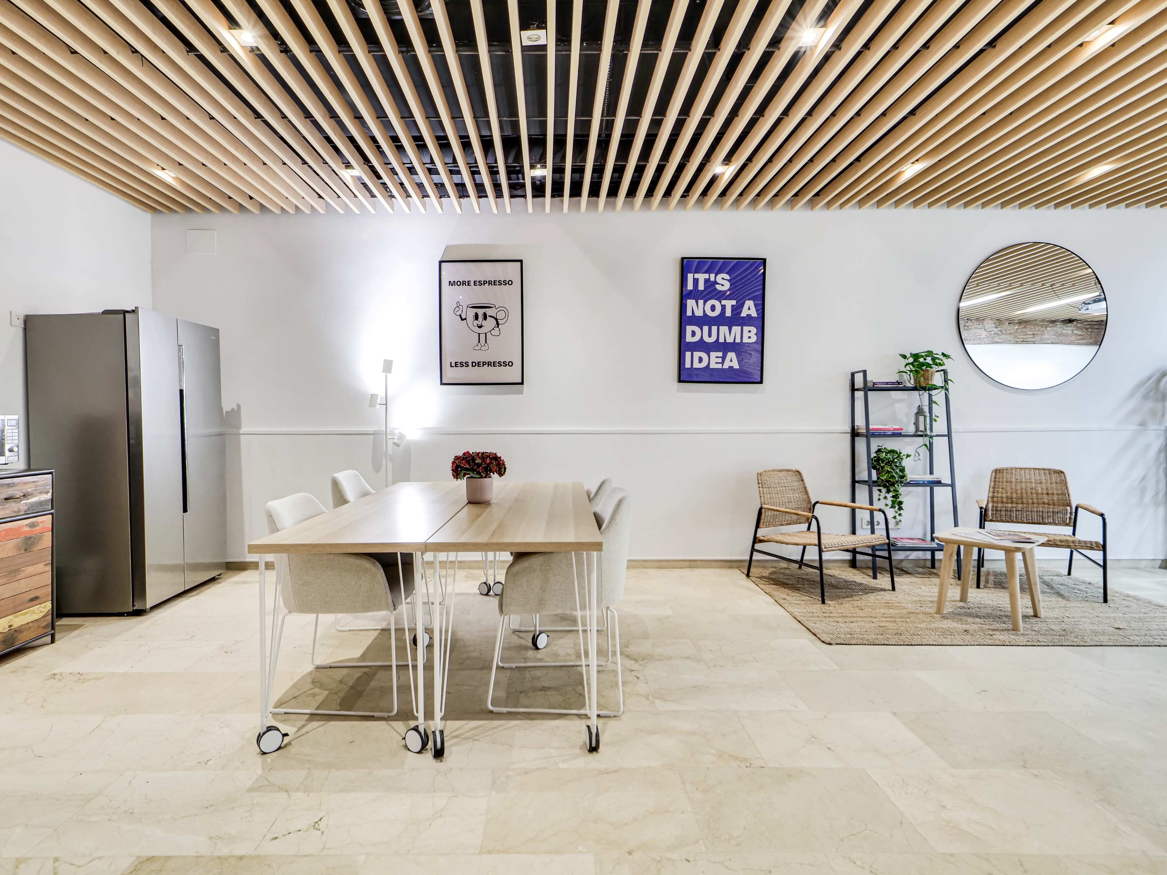 A modern office space features a wooden dining table with chairs, a kitchenette, and seating area with two chairs beside a circular mirror.