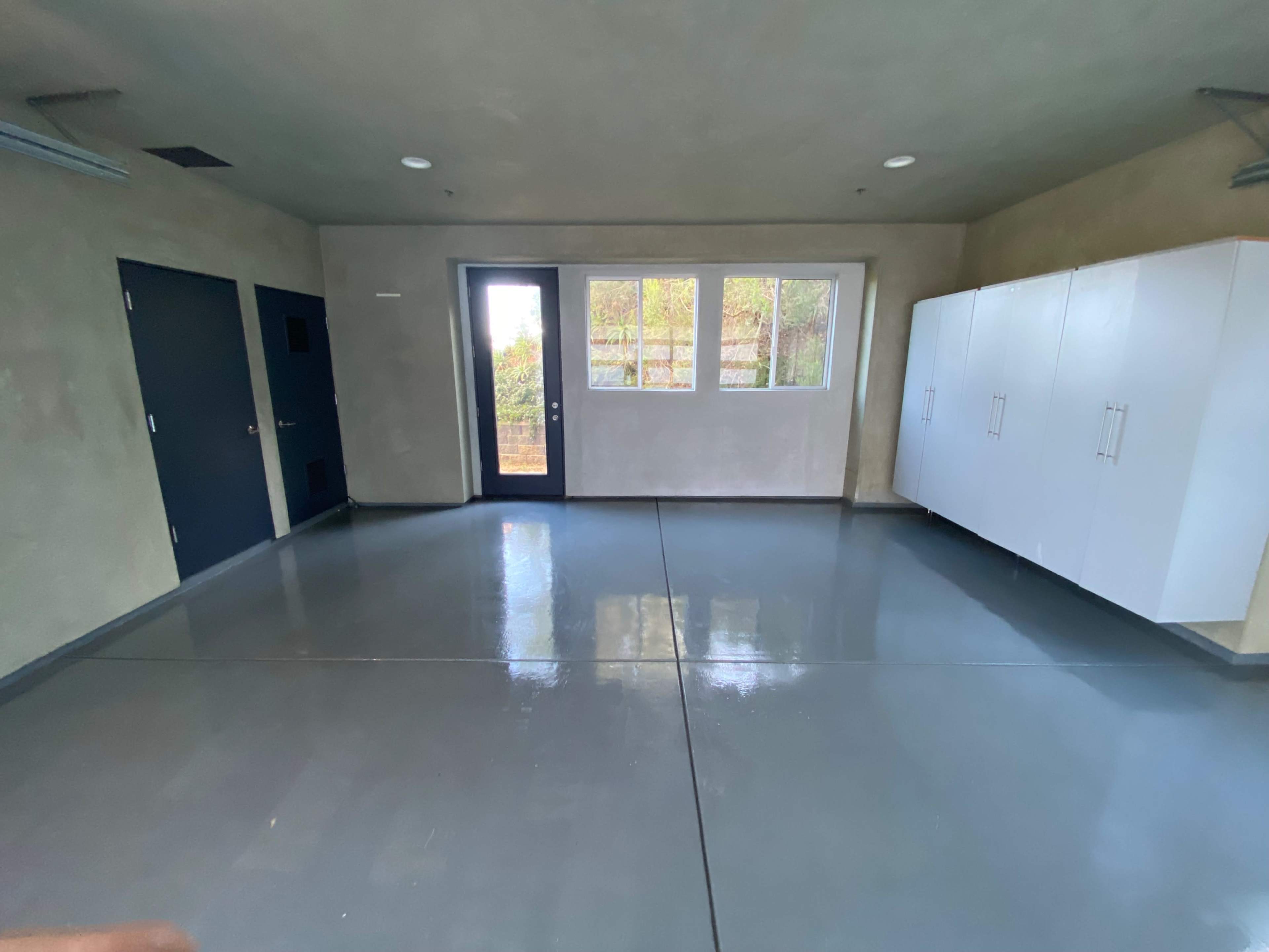 The image shows a vacant room with gray floors, a large window, a door, and white cabinets along one wall.