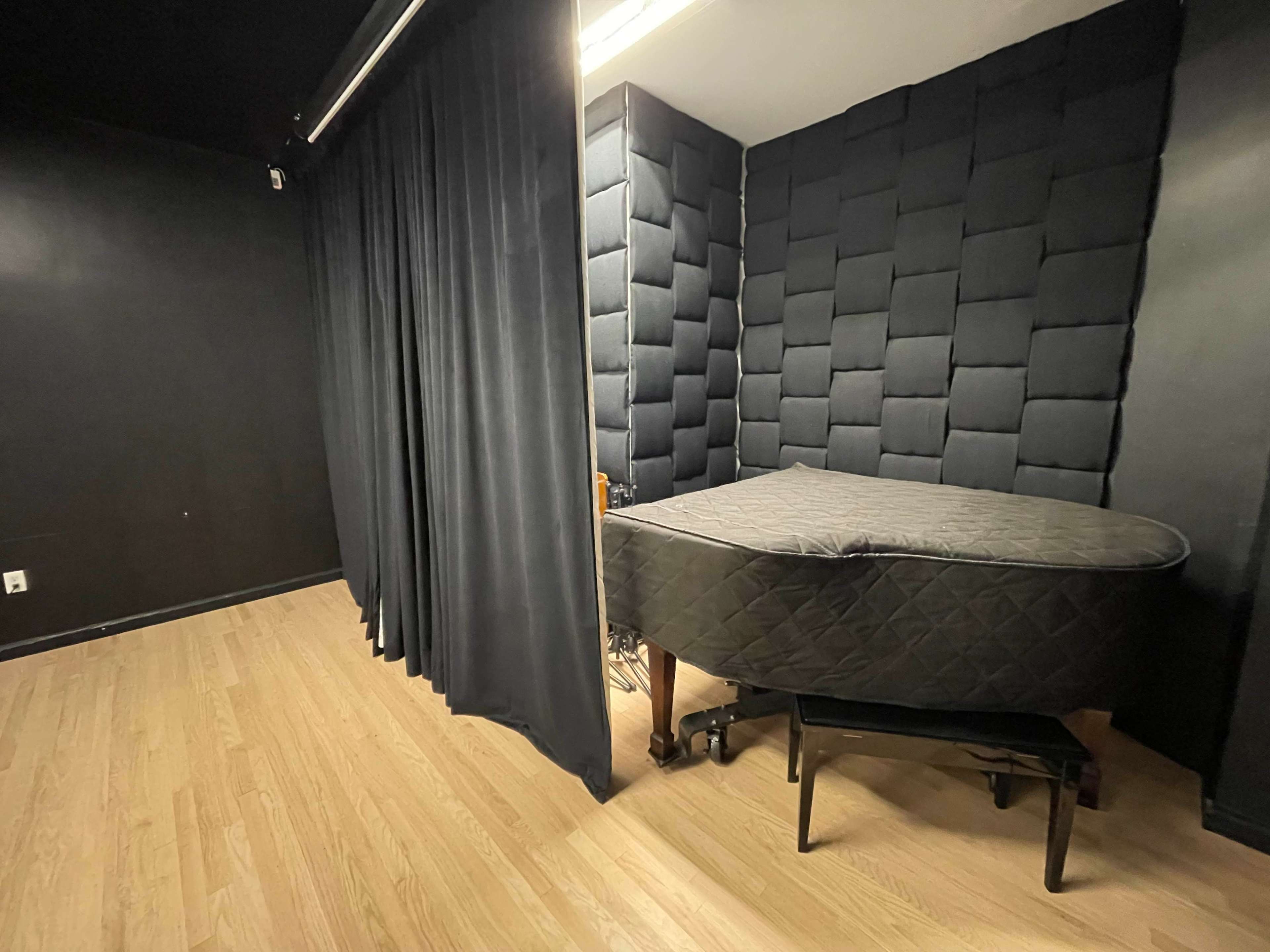 The image depicts a small room with a black, padded wall and a grand piano covered with a dark cloth, separated by a curtain from the rest of the space.
