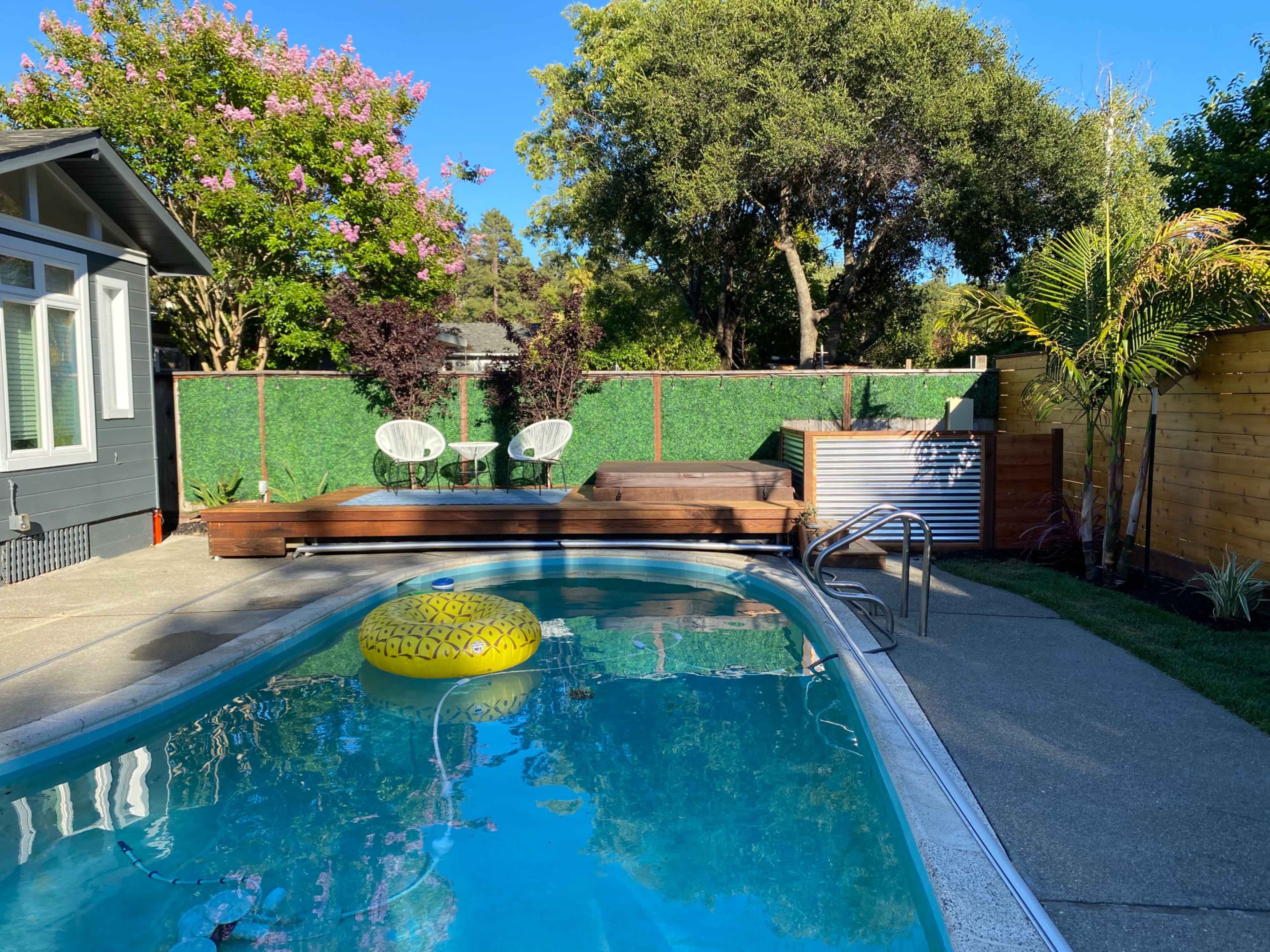 A backyard with a swimming pool, a wooden deck, a hot tub, and lounge chairs surrounded by greenery.