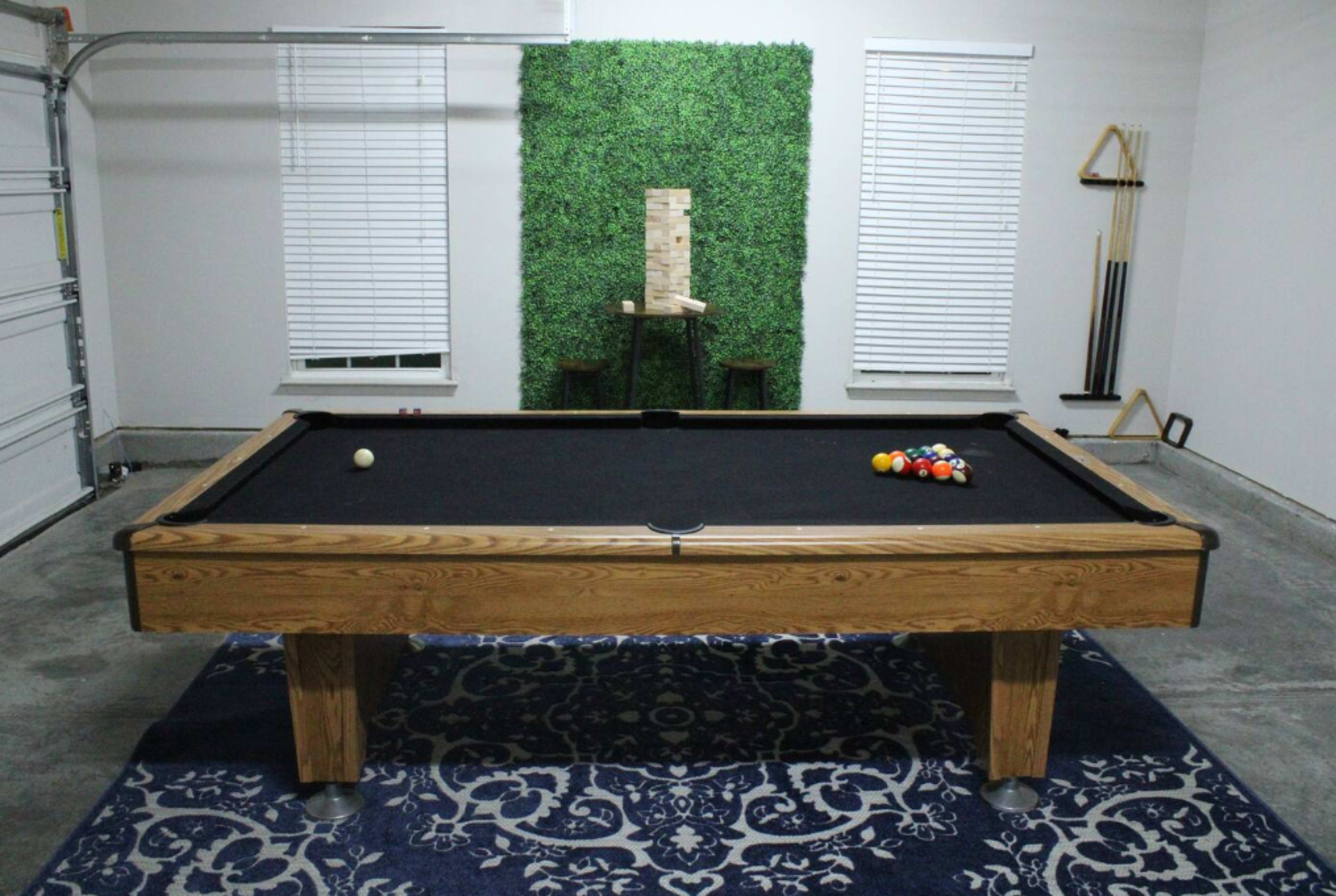A billiards table with colorful balls set up on it is positioned in a garage-like space featuring a green wall, wooden decor, and window blinds.