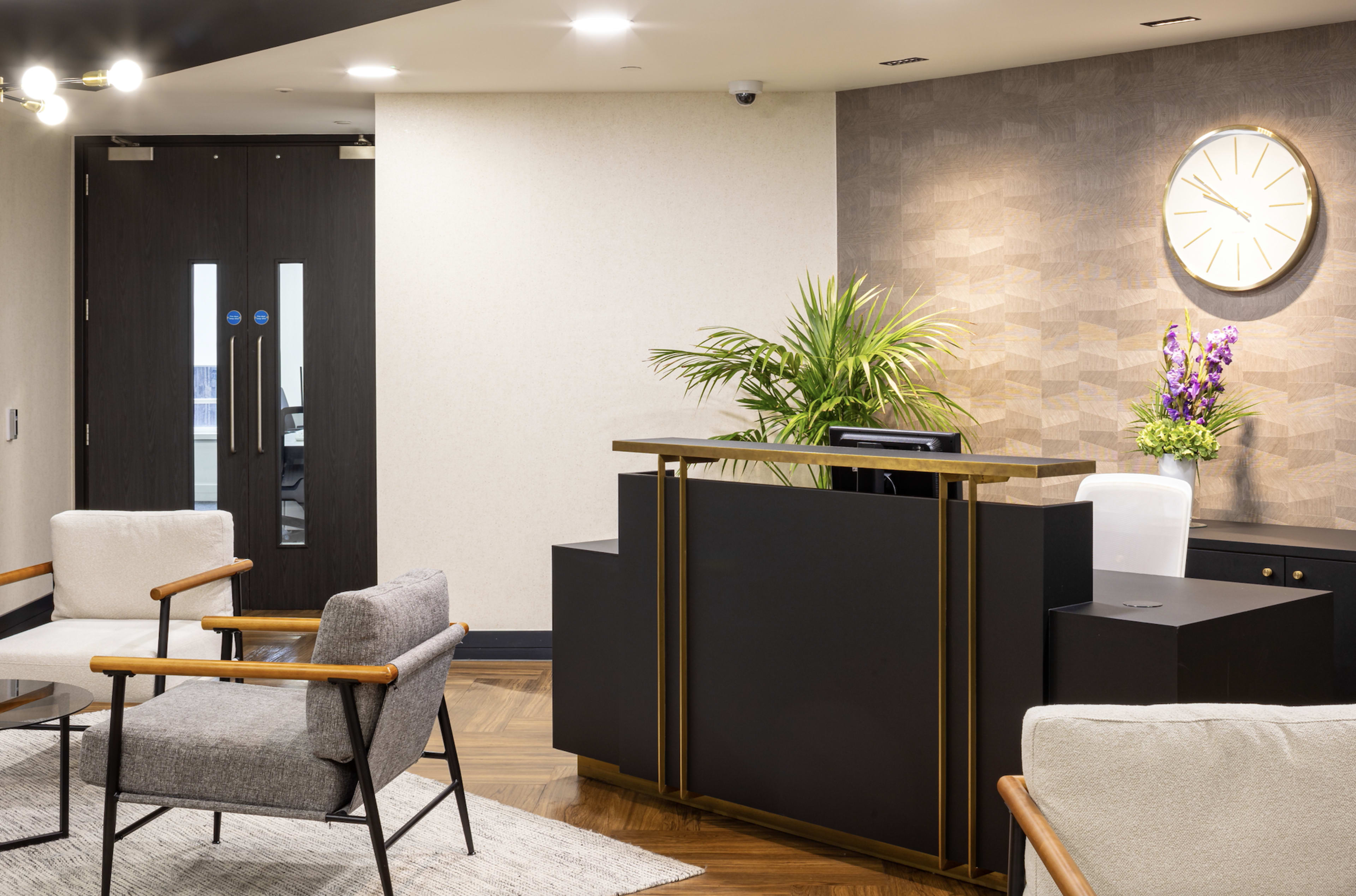 A modern reception area features a dark wooden front desk, a wall clock, and seating arrangements with armchairs and a rug.