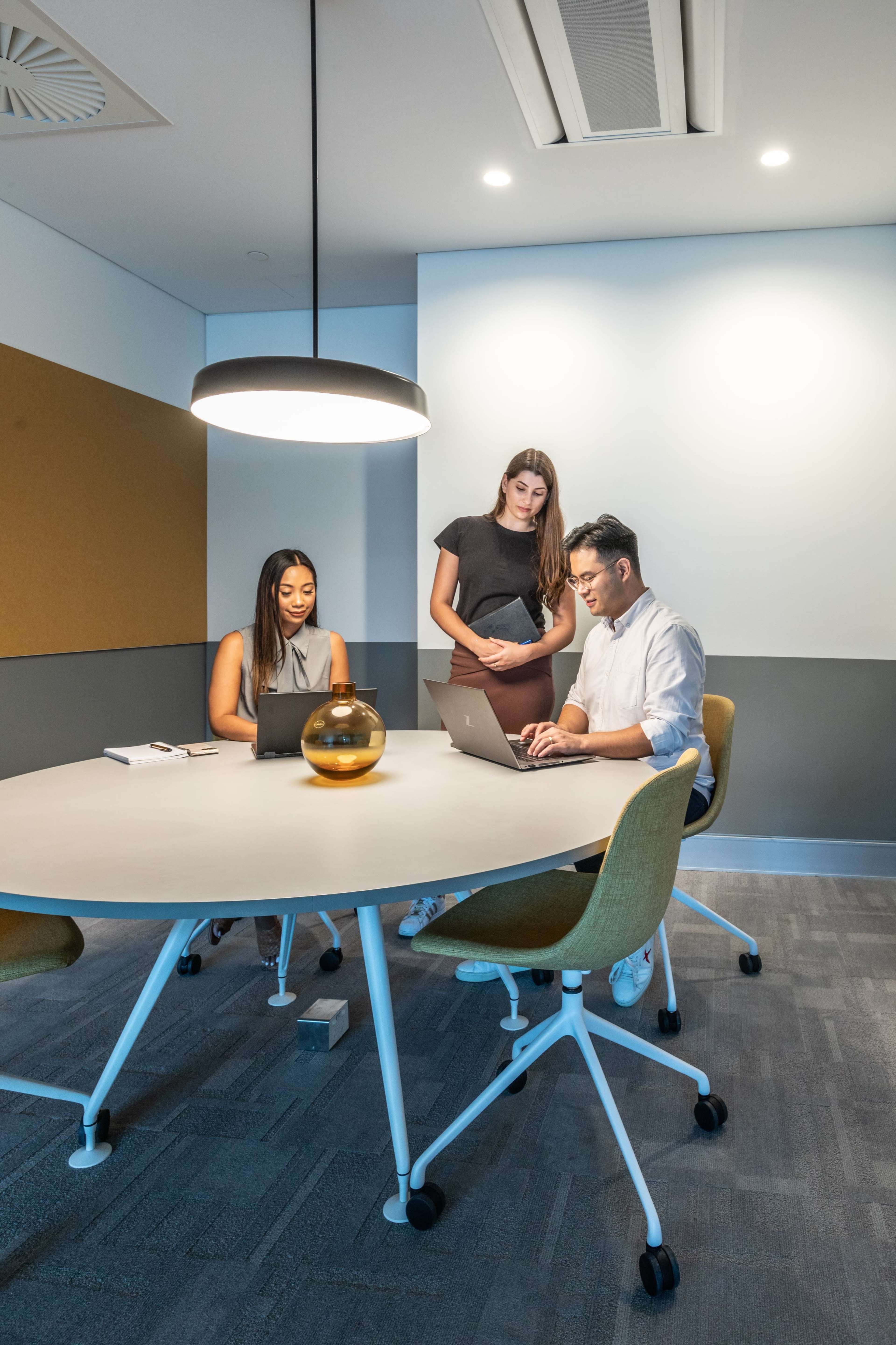 Three people are engaged in a discussion around a circular table in a modern meeting room, with laptops and a decorative glass object on the table.