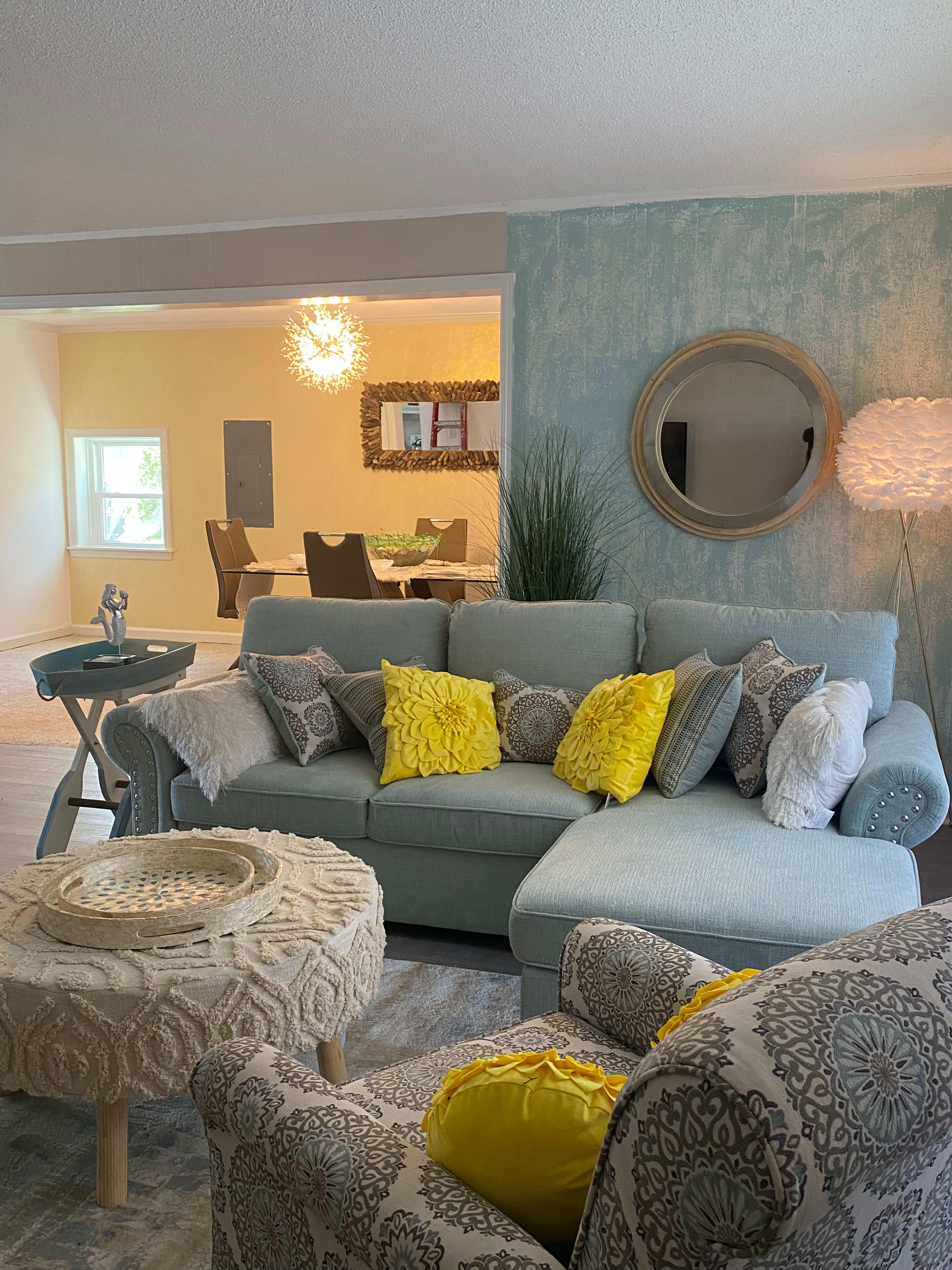 A light-colored living room features a sectional sofa with yellow and gray decorative pillows, a round coffee table, and a dining area visible through an archway.