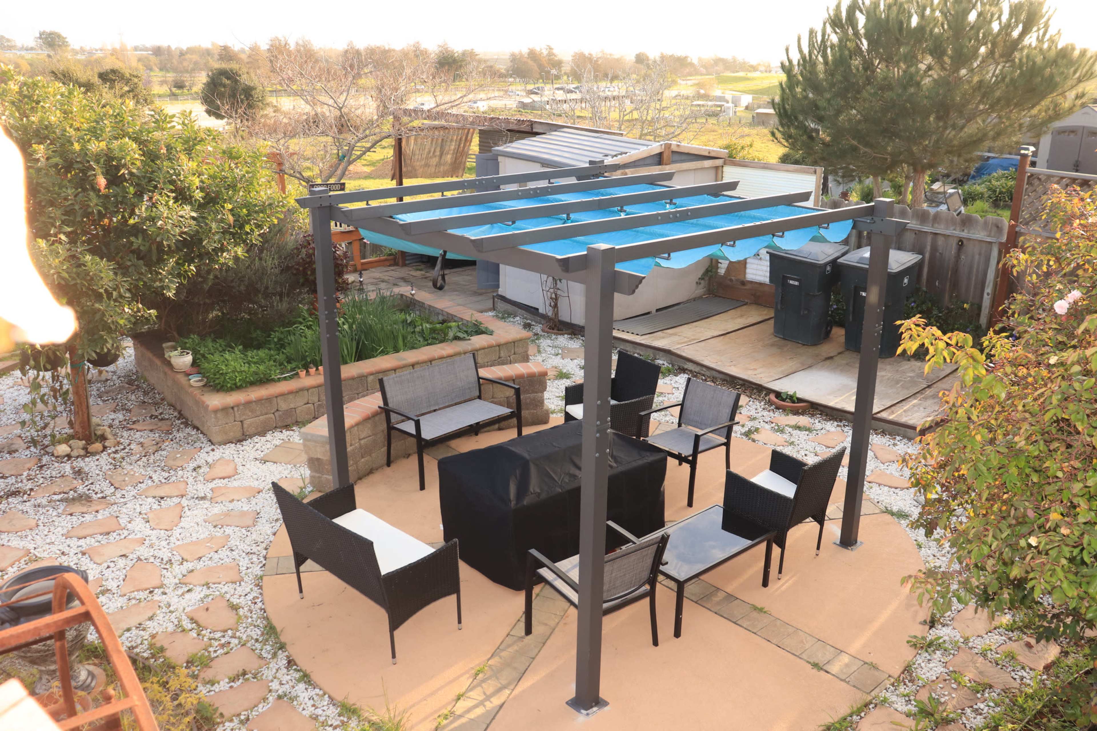 The image shows a patio area with a metal pergola, outdoor seating, and a square table in a landscaped yard.